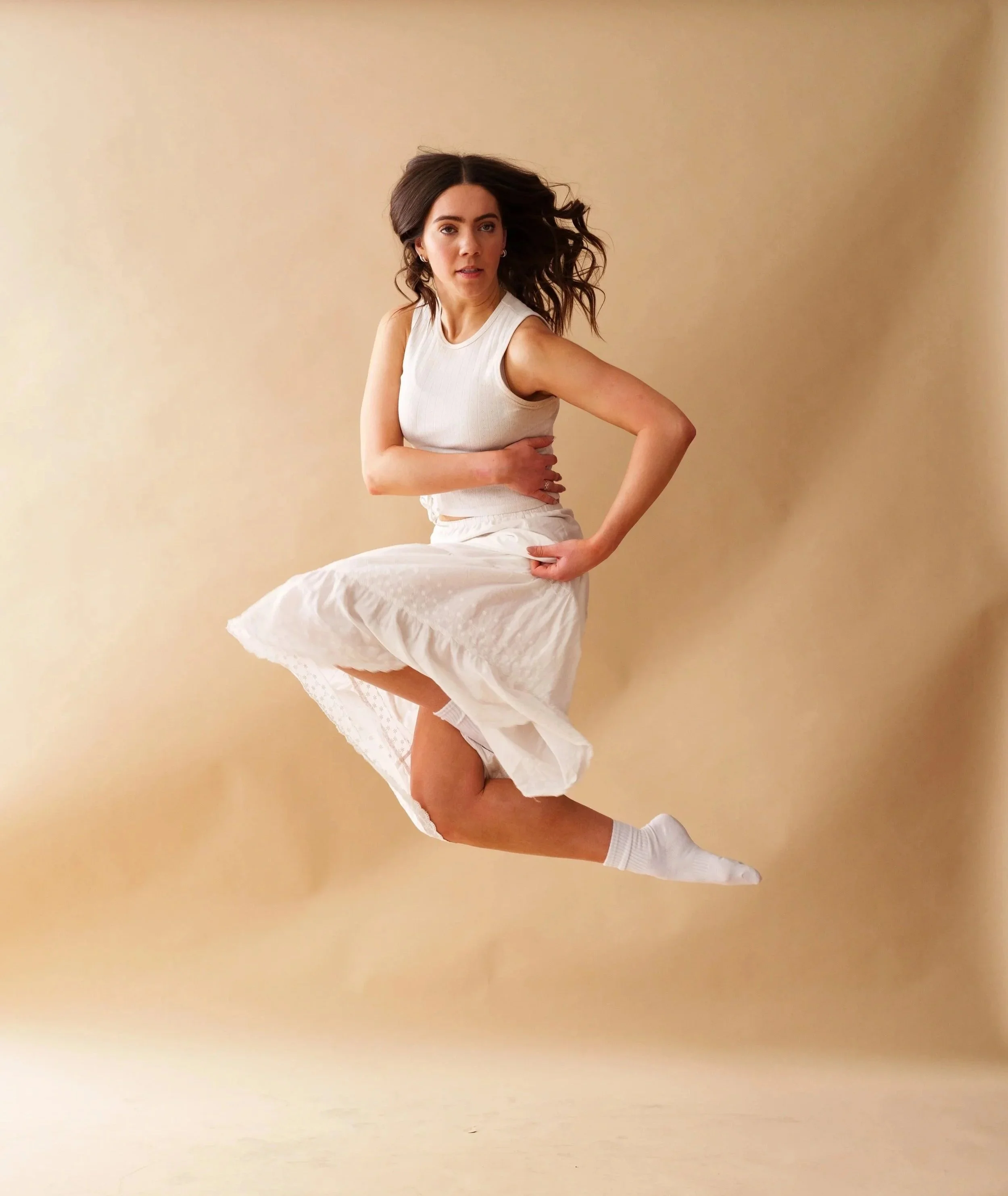A woman in a white sleeveless top and skirt is mid-air, jumping with one knee bent and her hair flowing, against a beige background.
