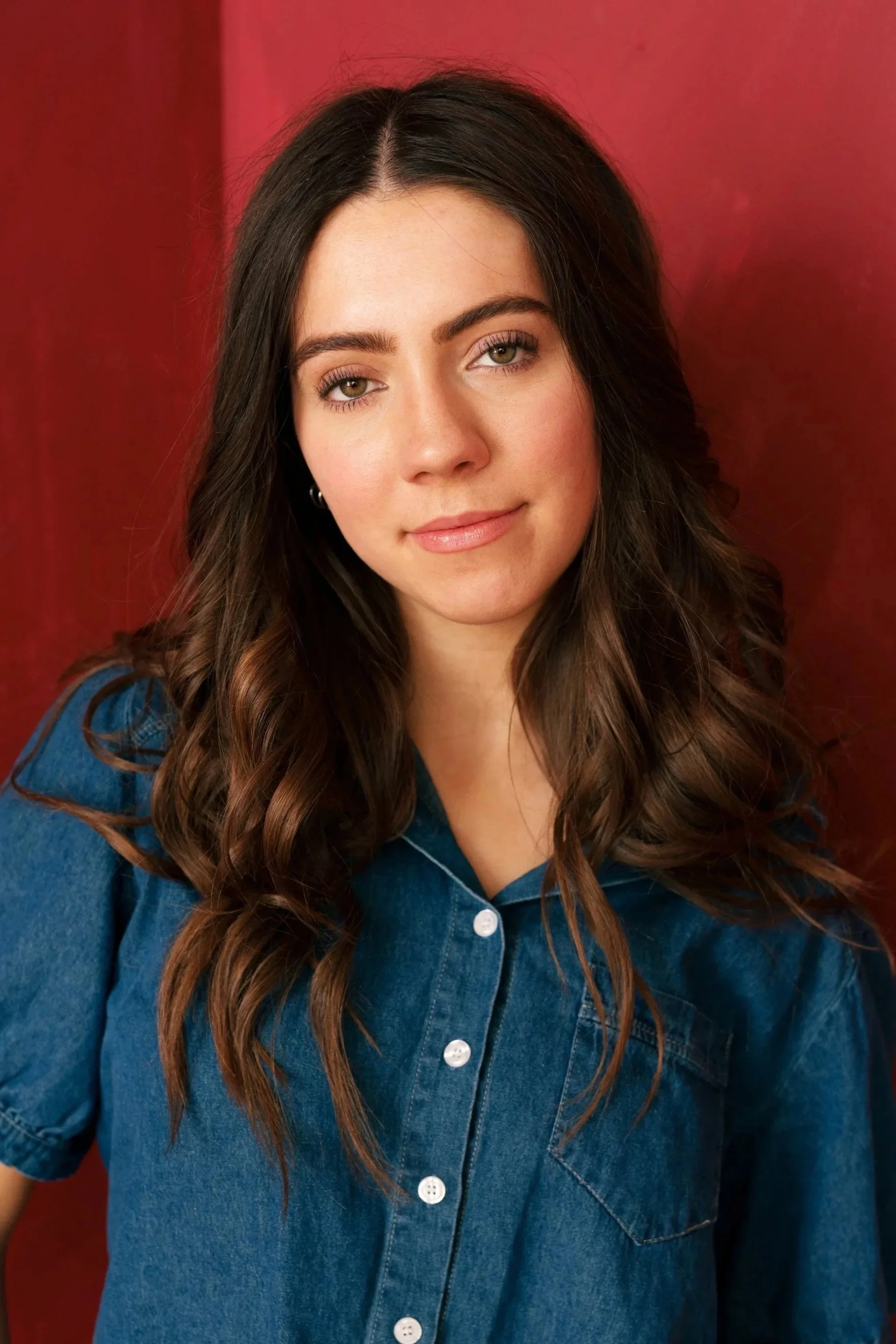 A young woman with wavy brown hair, wearing a blue denim shirt, standing against a red background.