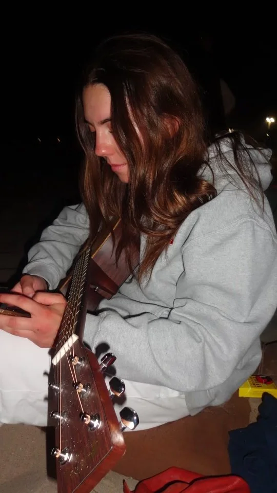 Young woman with brown hair wearing a gray hoodie, playing an acoustic guitar while looking at her phone outdoors at night.