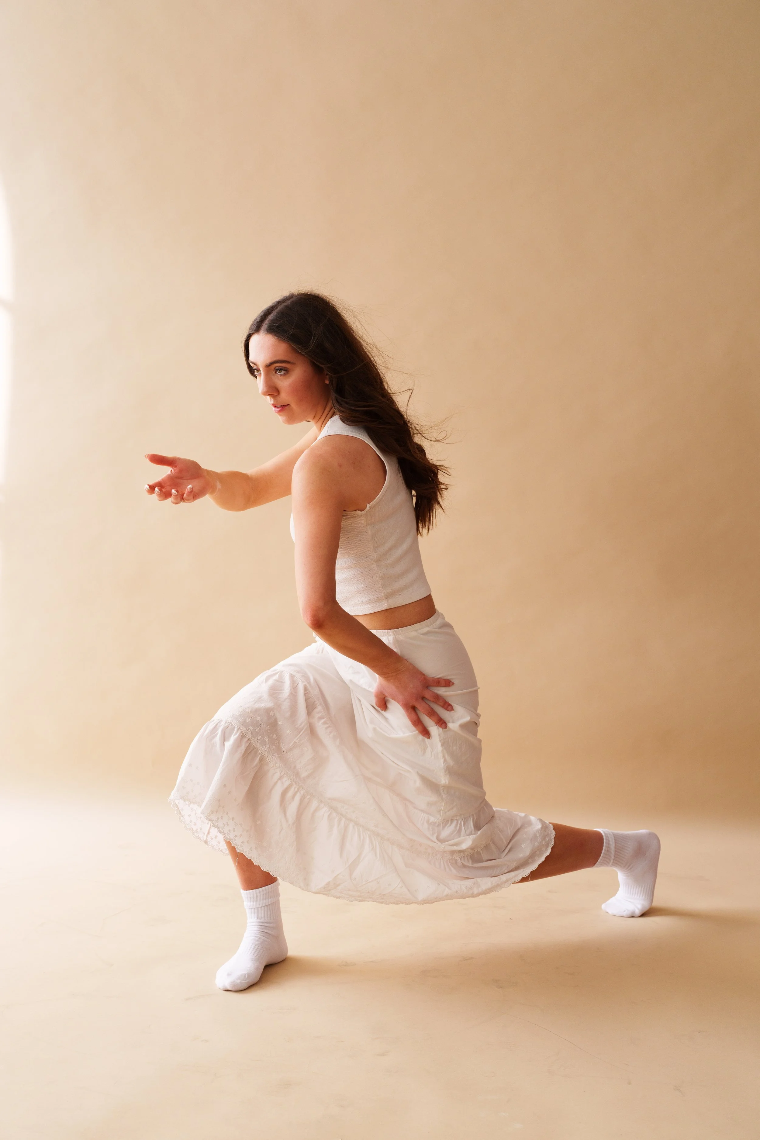 Young woman dancing or performing a movement in a neutral studio space, wearing a white crop top, long white skirt, and white socks, mid-movement with one arm extended and the other hand on her hip.