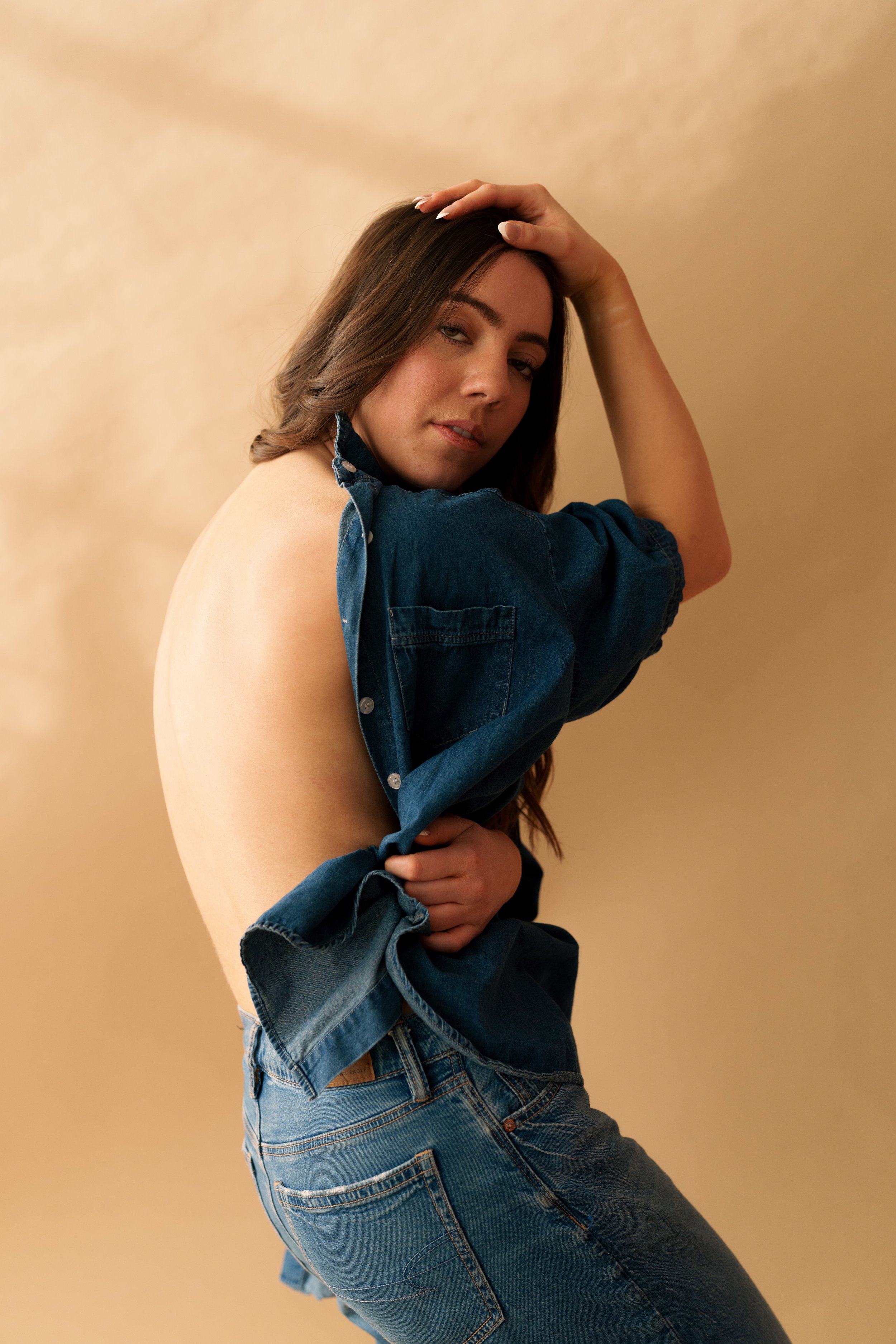 A woman with long brown hair poses against a beige background, wearing a partially open denim shirt and jeans.