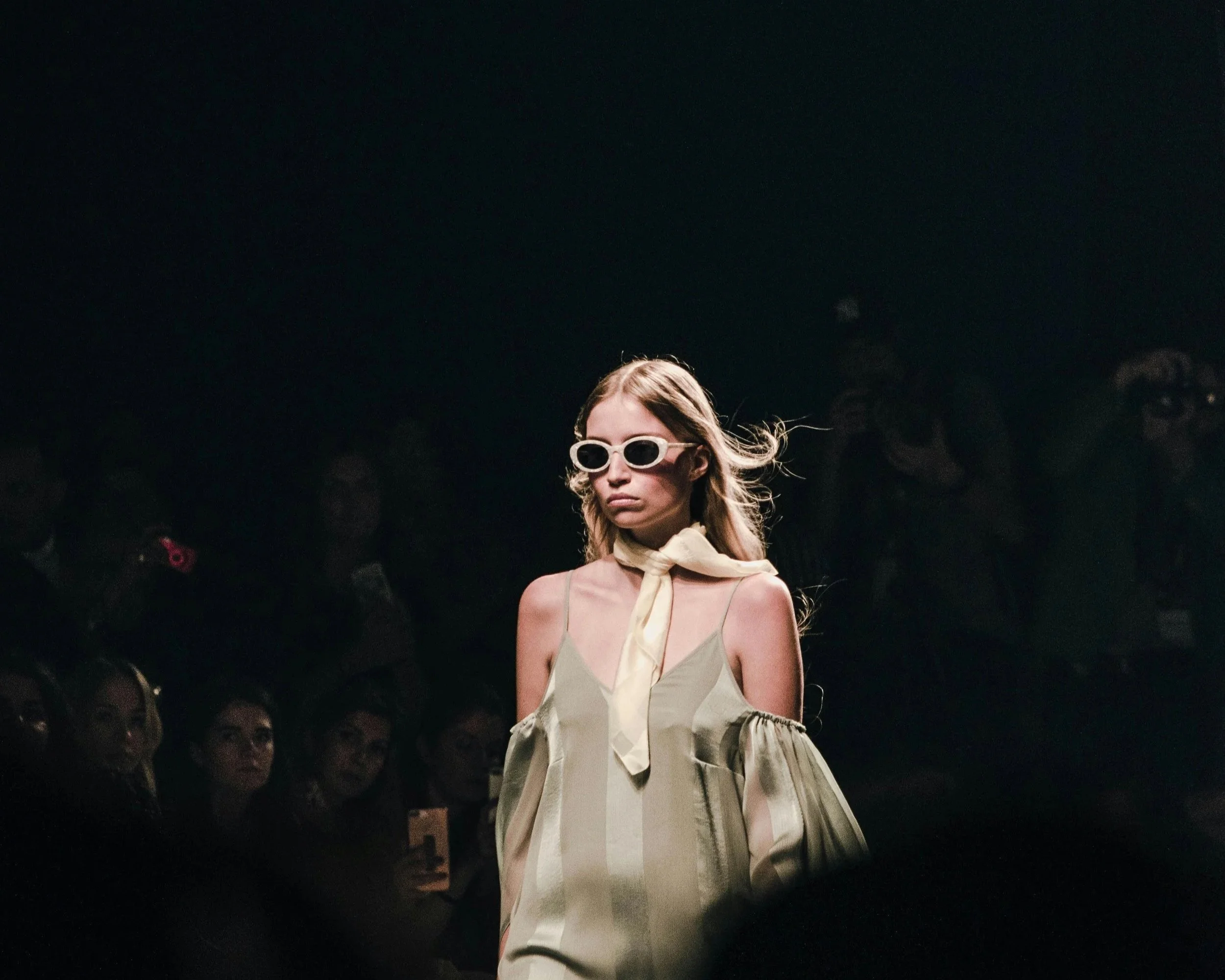 Fashion model walking on runway wearing beige and pastel-colored dress with a large bow, sunglasses, and belt, with audience watching in dark background.