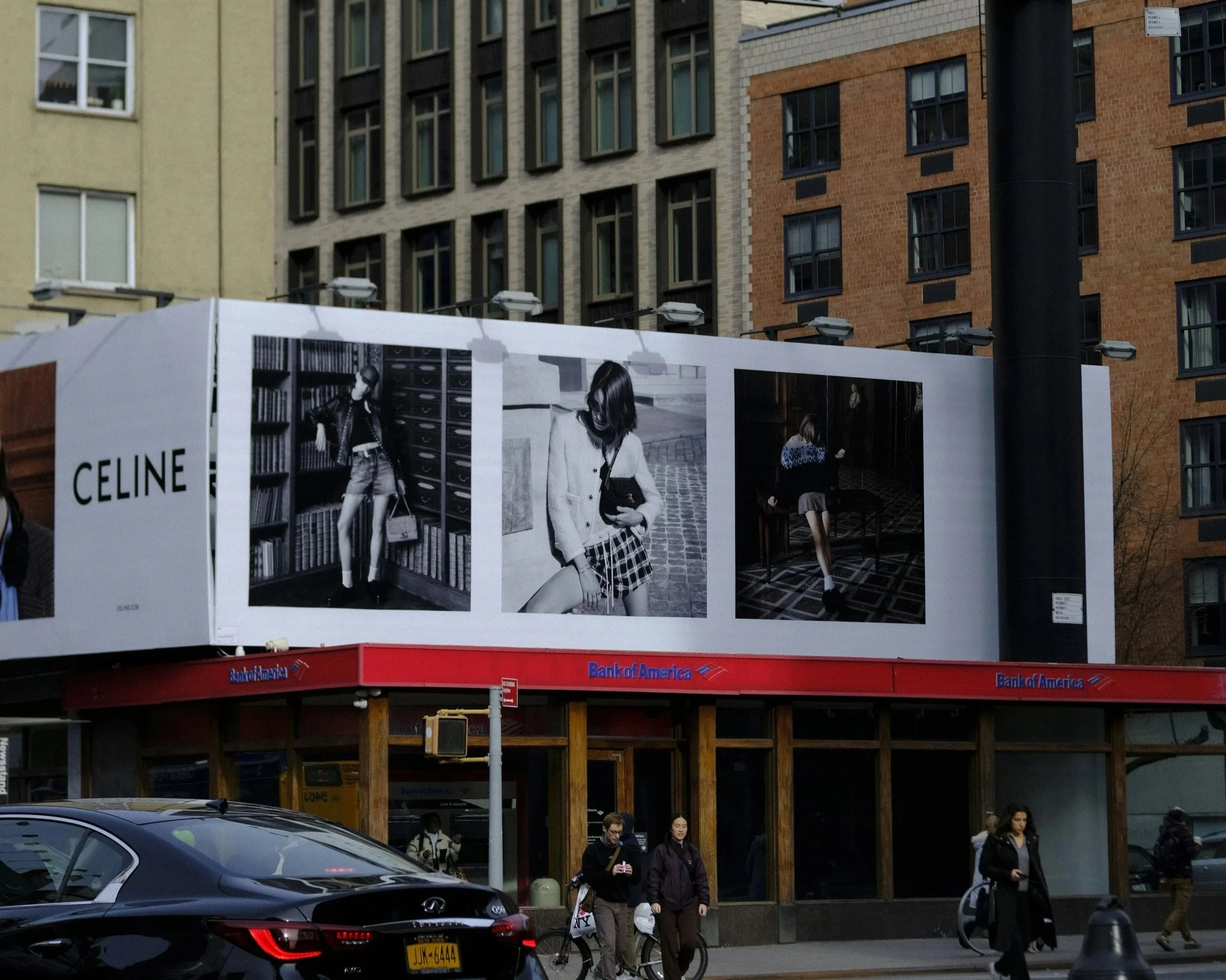 Street scene with pedestrians, parked cars, and indoor fashion photos displayed on a billboard outside a building with a Bank of America storefront.