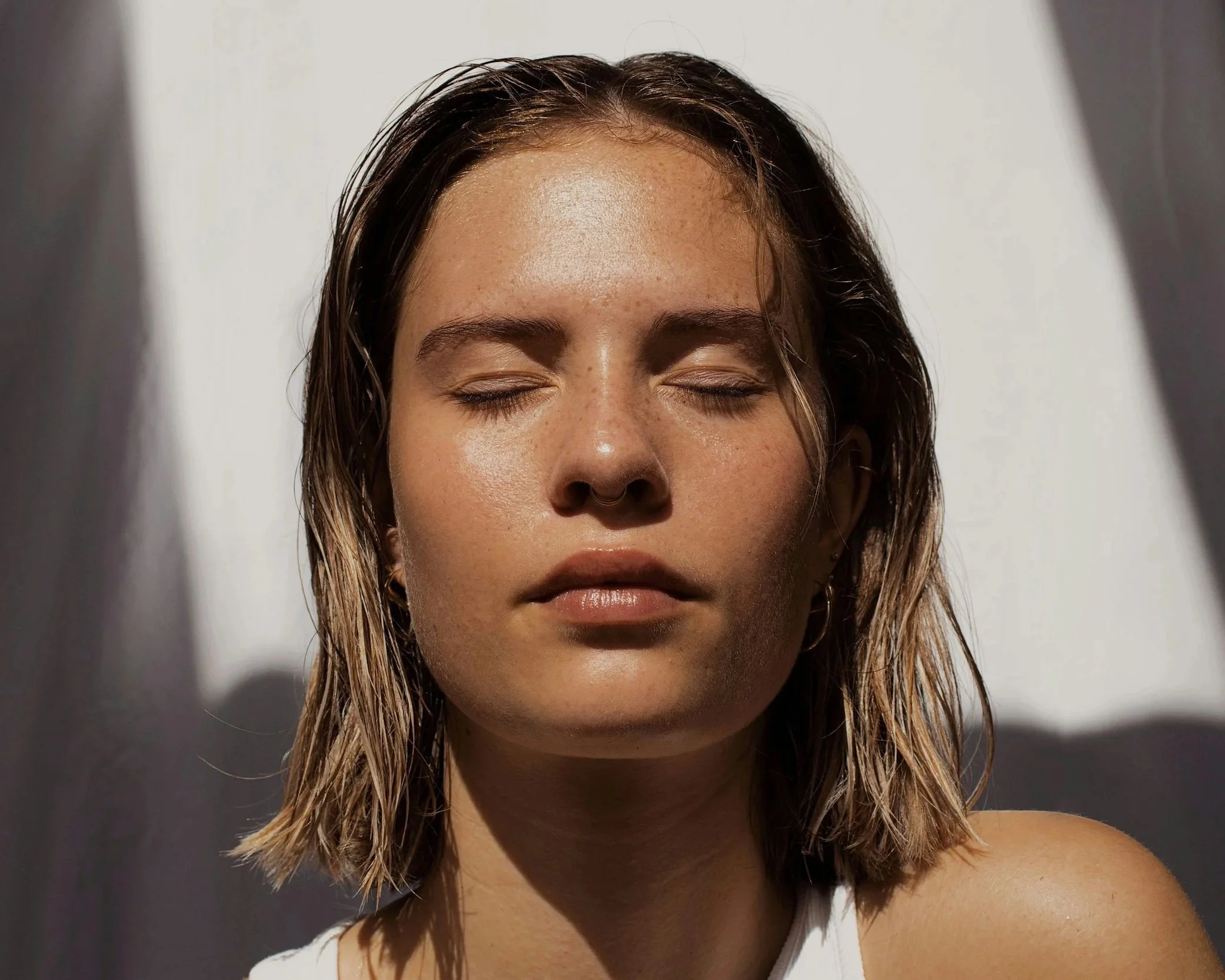 Close-up of a woman's face with eyes closed, wet hair, and natural skin with freckles, lit by sunlight.