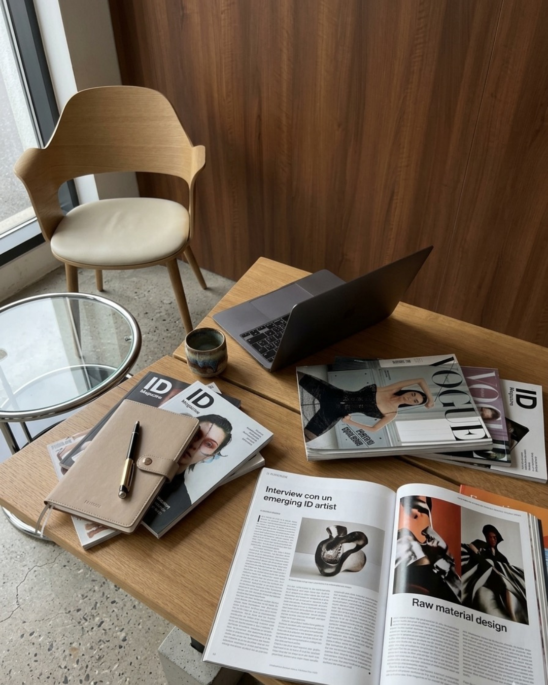 A wooden desk with an open laptop, magazines, a notebook, pen, and a ceramic mug. An empty wooden chair and a small round glass-top side table are nearby, with a window in the background.