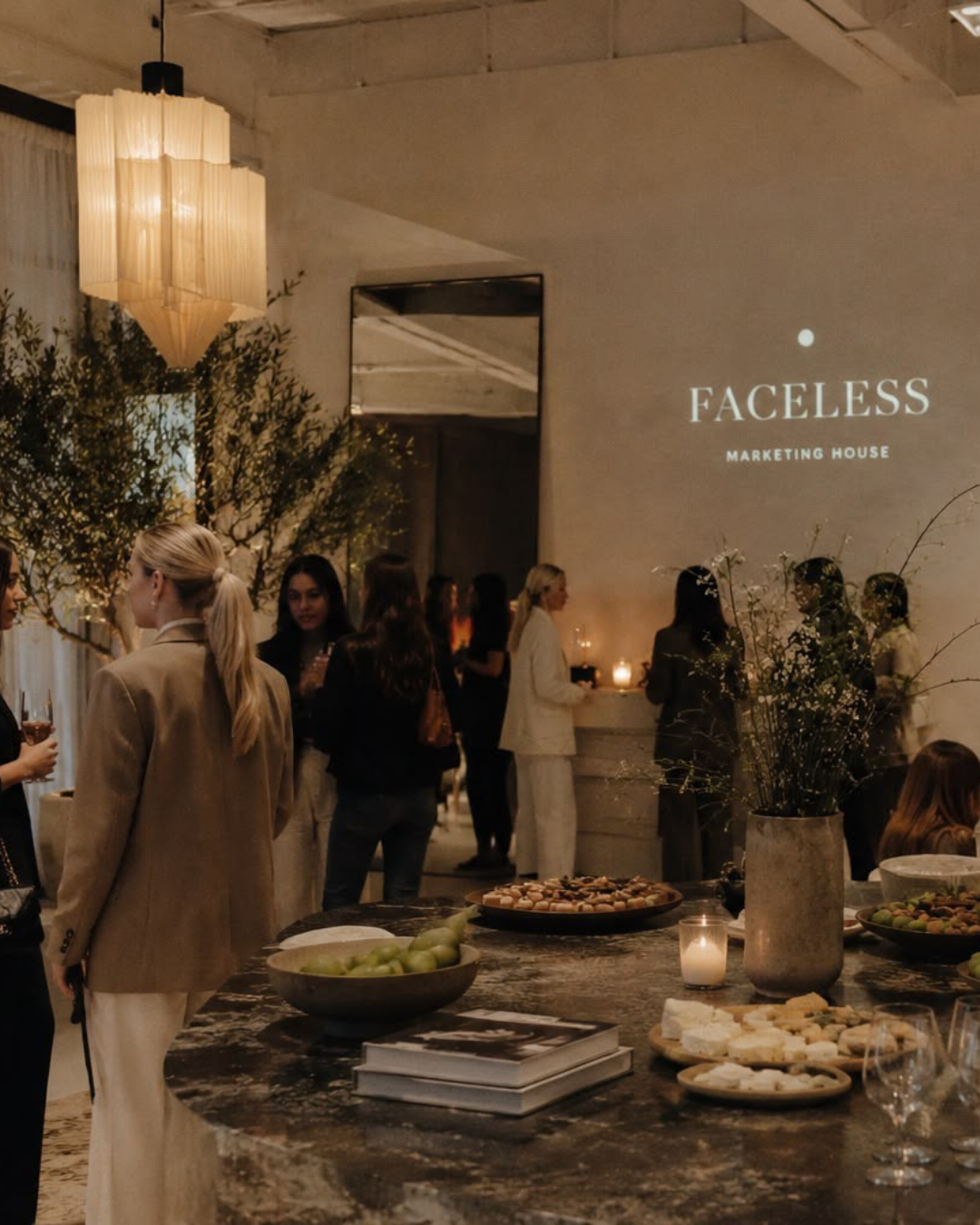 Elegant indoor event with people socializing around a marble table, floral arrangements, and a projection on the wall reading 'FACELSS Marketing House'.