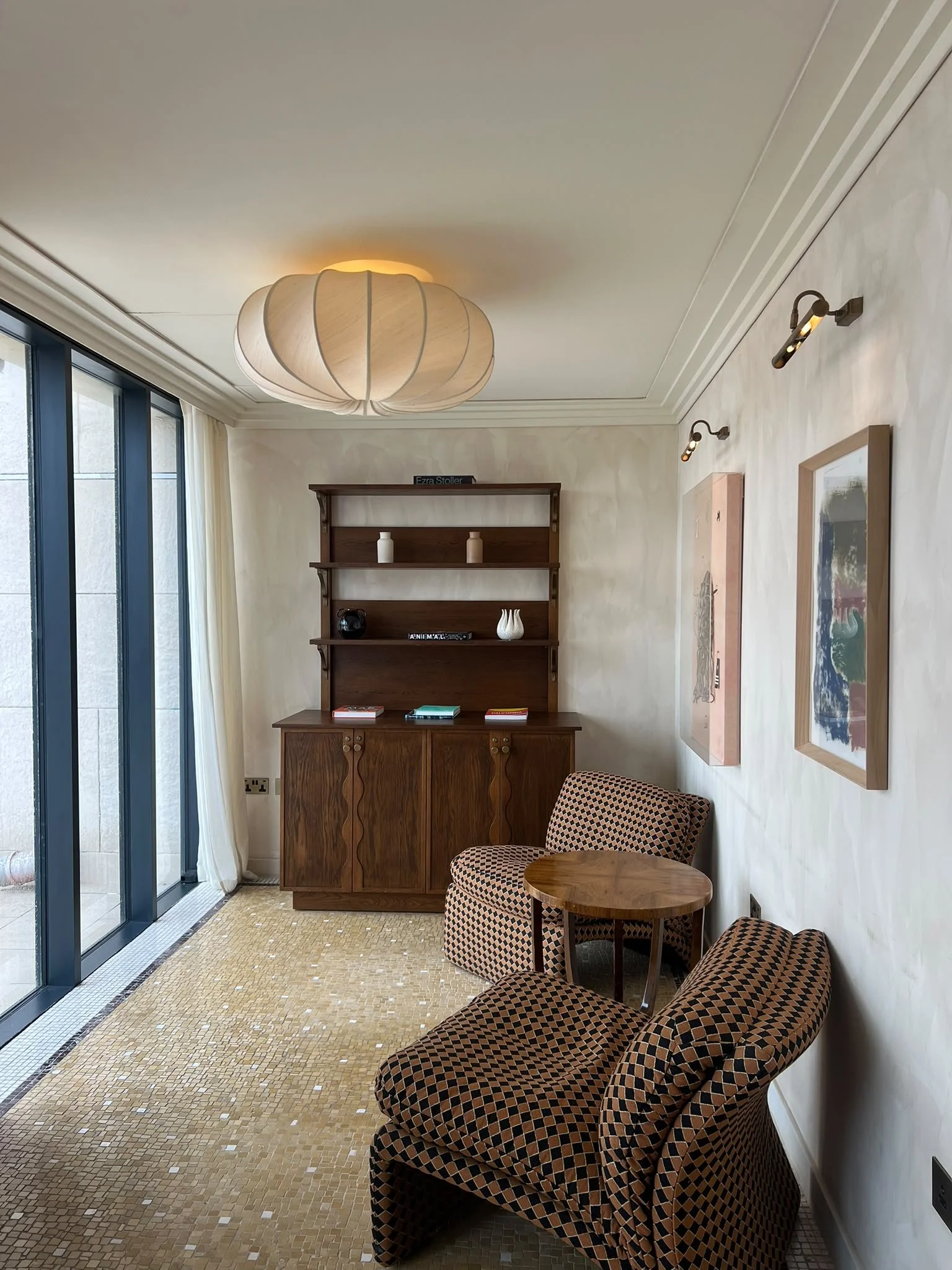 Interior of a room with a beige patterned carpet, a wooden cabinet with decorative items, a small round wooden table, and upholstered chairs with a geometric black and brown pattern. Large floor-to-ceiling windows with white curtains, a framed artwork on the wall, and modern wall-mounted lights.