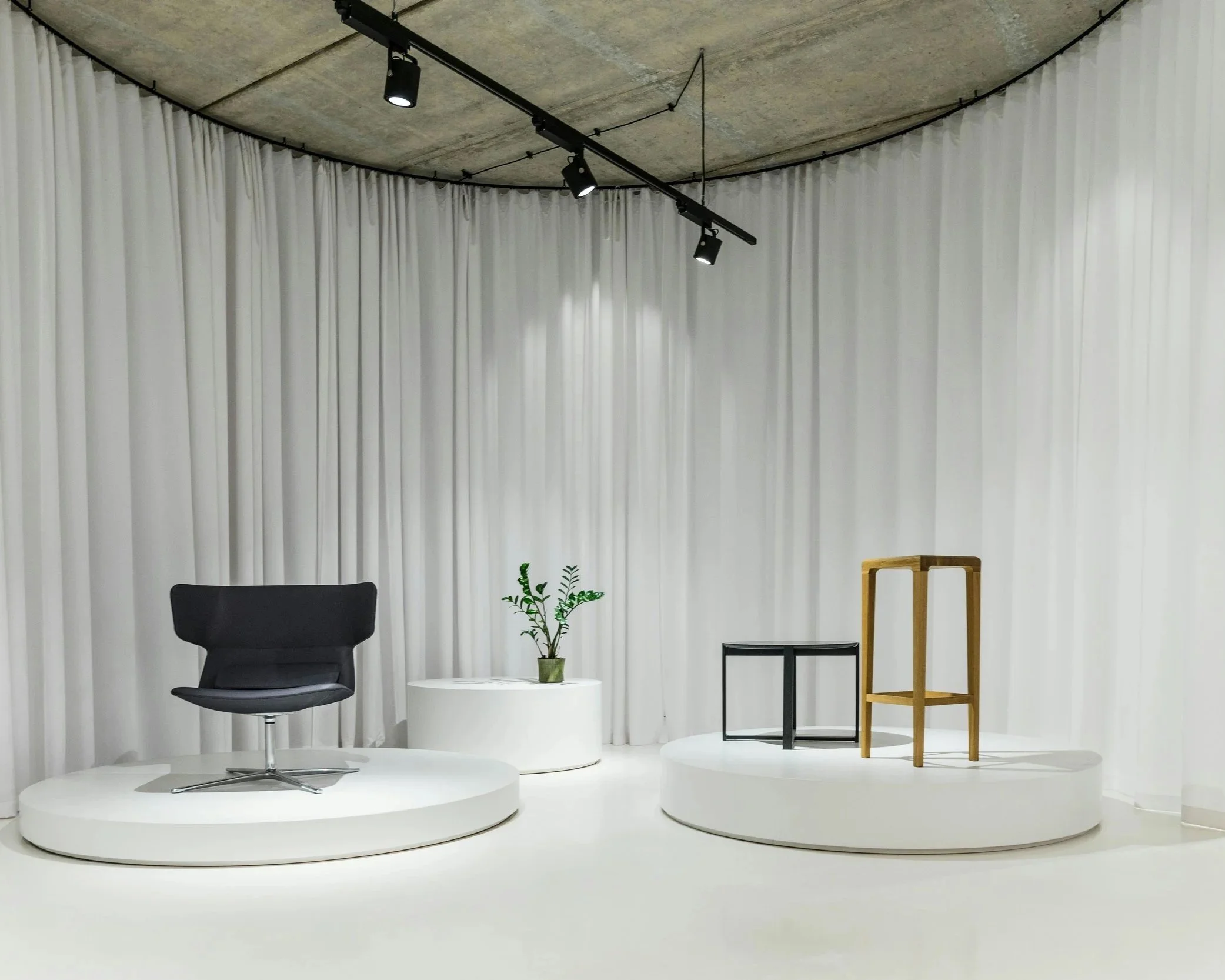 A stage setup with a black swivel chair on a white circular platform, a white round table with a small green potted plant, a tall wooden chair, and a modern black metal table with a geometric design, all against a background of white curtains and a concrete ceiling with black track lighting.