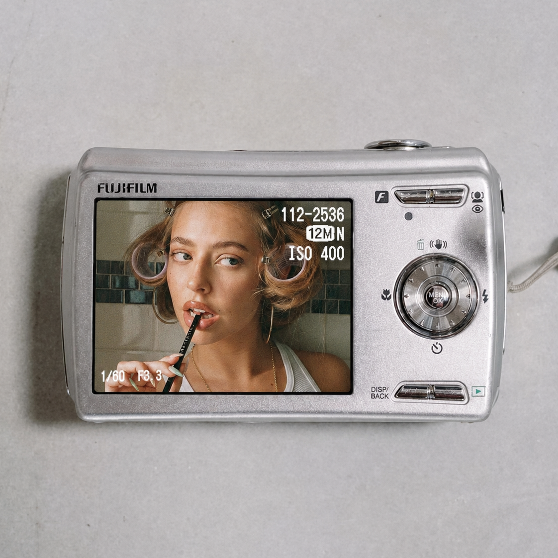 A silver compact digital camera displaying a selfie of a woman with curlers in her hair, lipstick, and hoop earrings, holding a makeup brush near her lips.