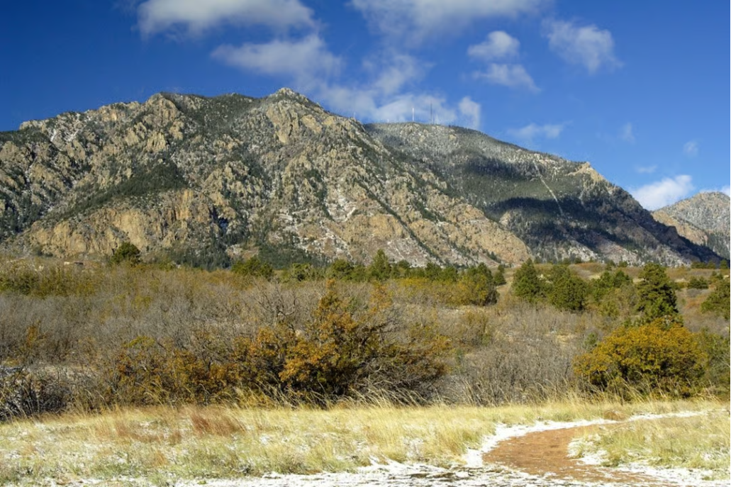 Scenic view of a Cheyenne Mountain with a hiking trail, trees, bushes, and a partly cloudy blue sky.
