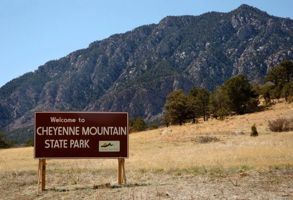 A sign that reads 'Welcome to Cheyenne Mountain State Park' in front of a mountainous landscape with trees and dry grass.