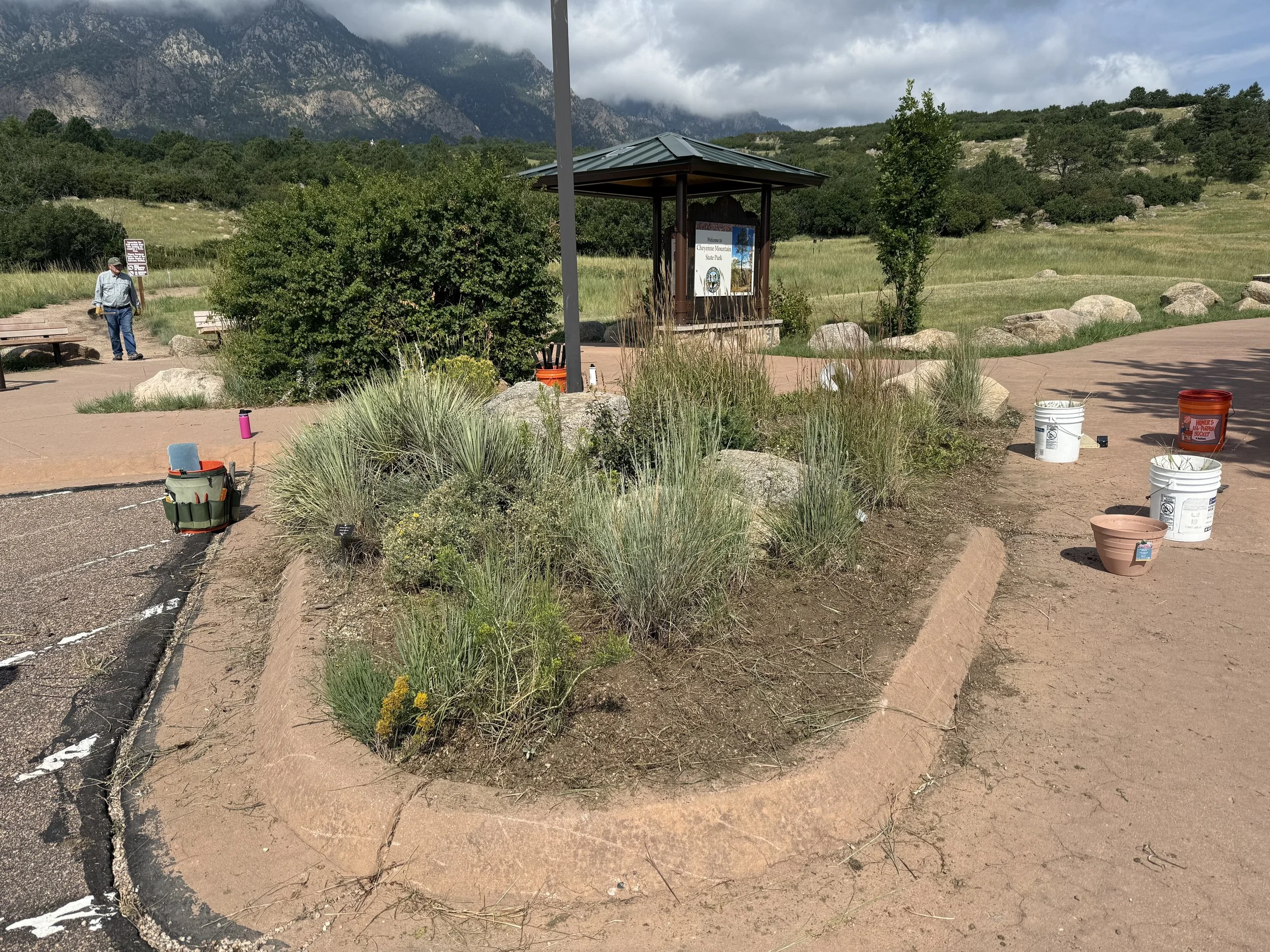 A scenic outdoor park with a sign for Cheyenne Mountain State Park, surrounded by greenery, rocks, and mountains in the background. There are buckets and tools indicating ongoing maintenance or landscaping work.