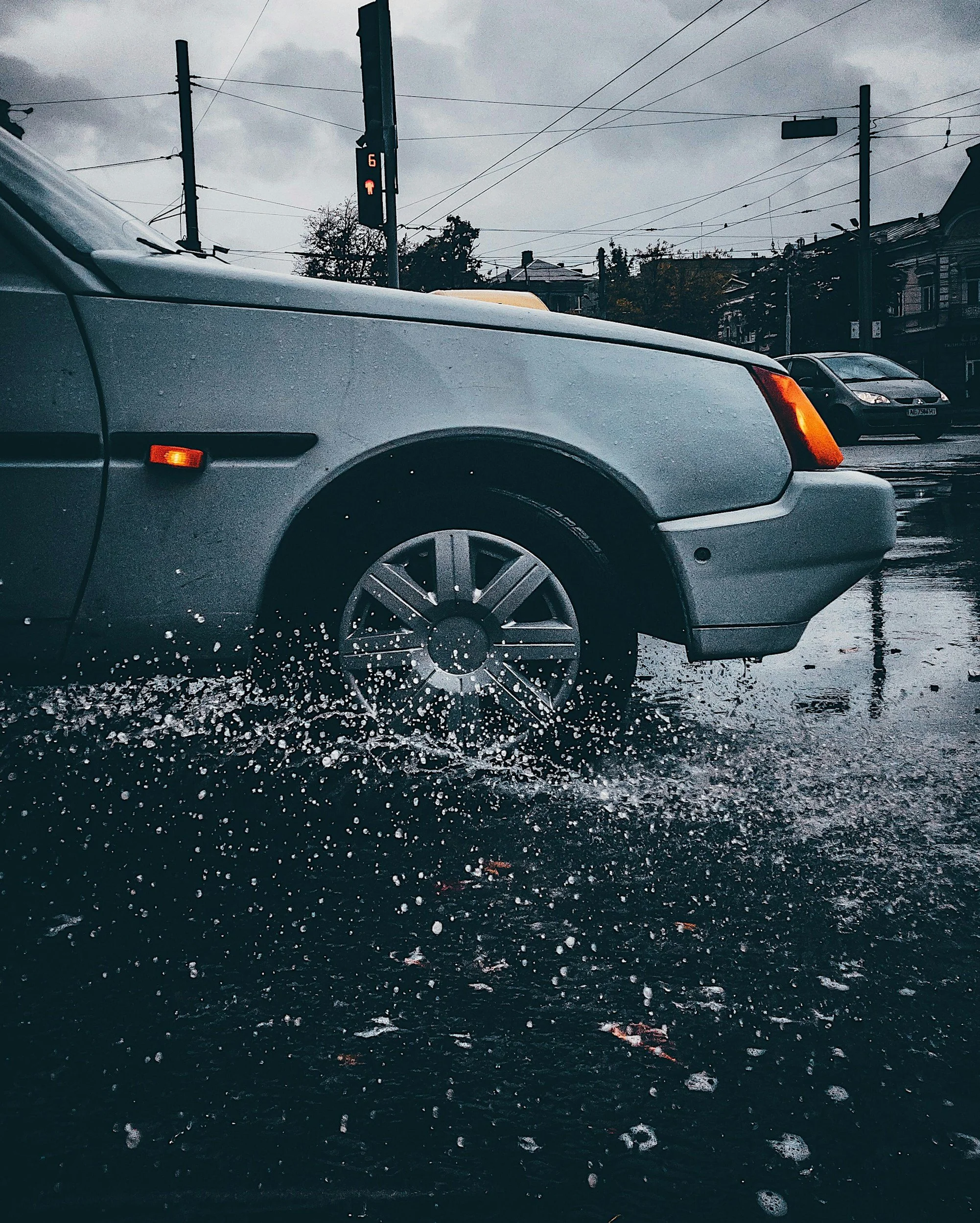 A car driving through a flooded street on a rainy day, with water splashing around the front tire.