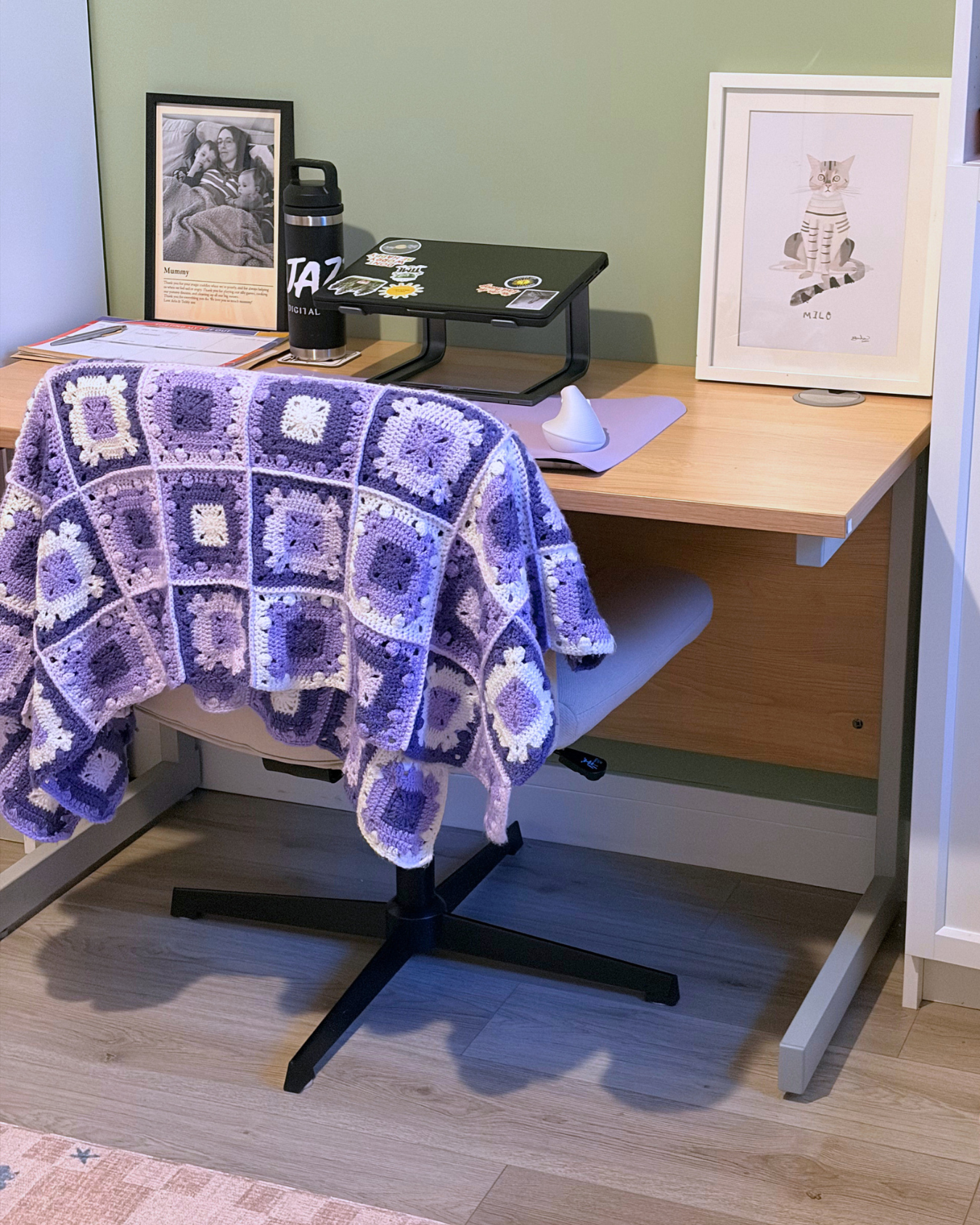 A tidy workspace with a wooden desk,office chair with a purple and white crocheted blanket draped over it, a framed photo of a woman and children, a small framed drawing of a cat named Milo, a black water bottle, a stack of papers, a laptop with stickers, a white mouse, and a purple silicone wrist rest.
