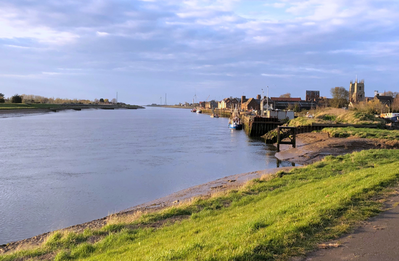 A riverside scene with a calm waterway, boats moored along the bank, grassy and rocky shoreline, and a village with a church tower in the background.
