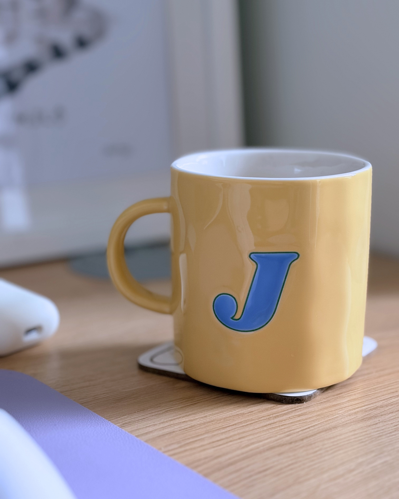 A yellow coffee mug with a blue letter 'J' on it sitting on a coaster on a wooden desk.