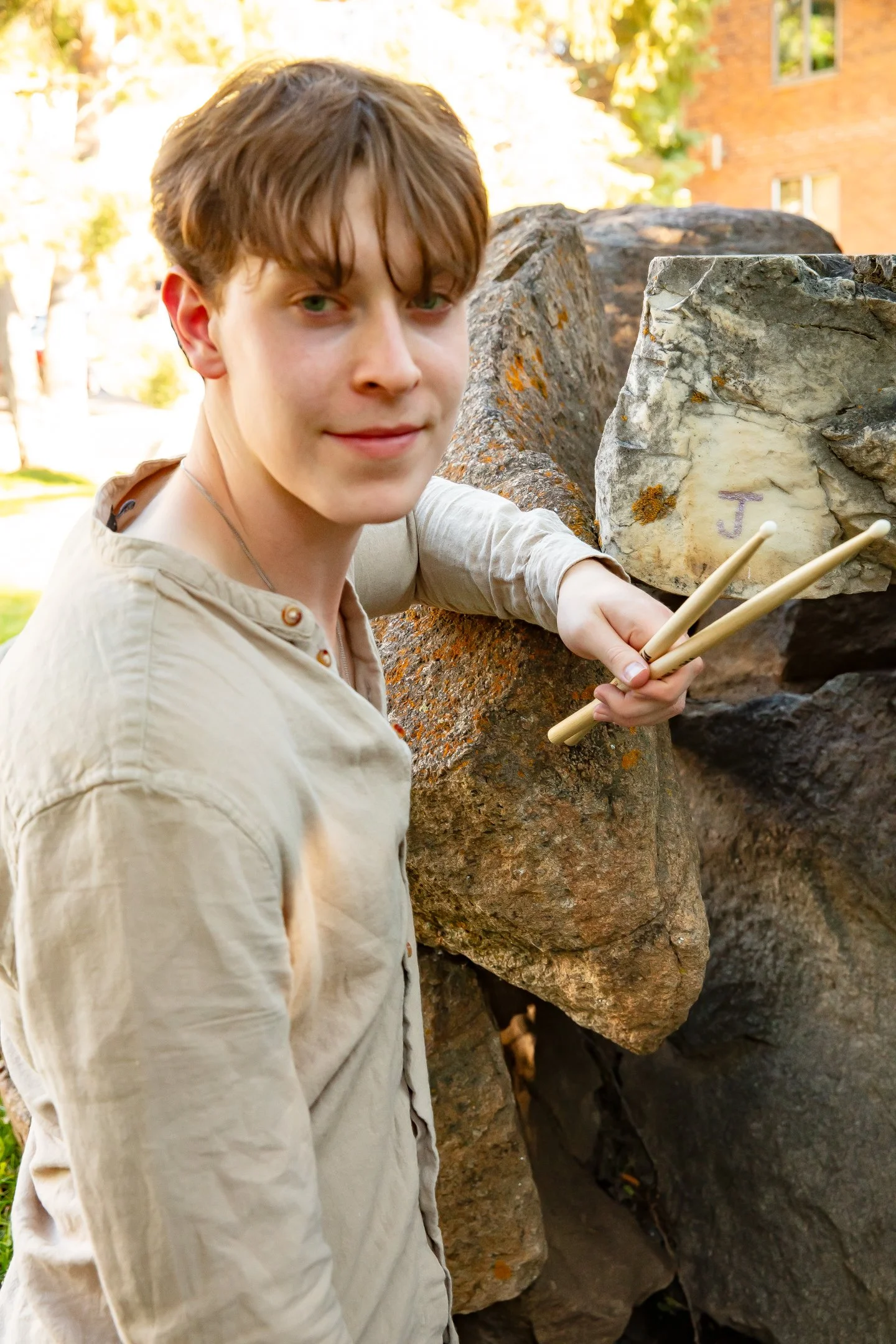 Young man holding drumsticks, leaning on rocks in a park.