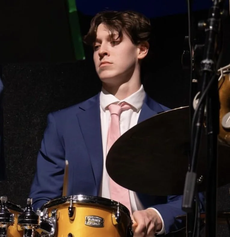 Young man in a blue suit and pink tie playing the drums.