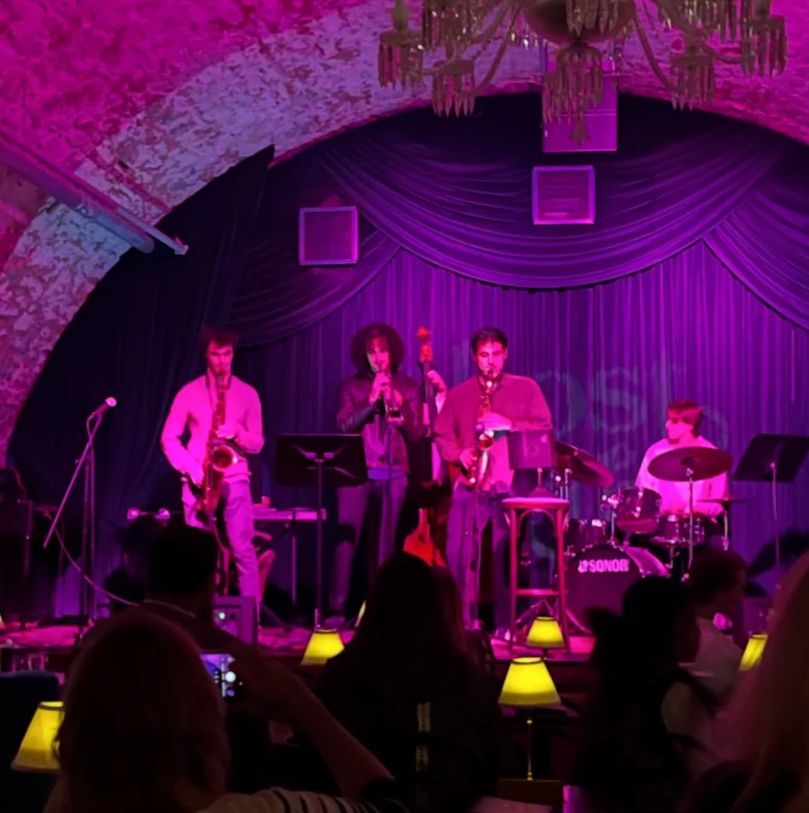 A jazz band performing on stage under pink and purple lighting in a cozy venue with a brick ceiling and chandelier, with audience members seated at tables.