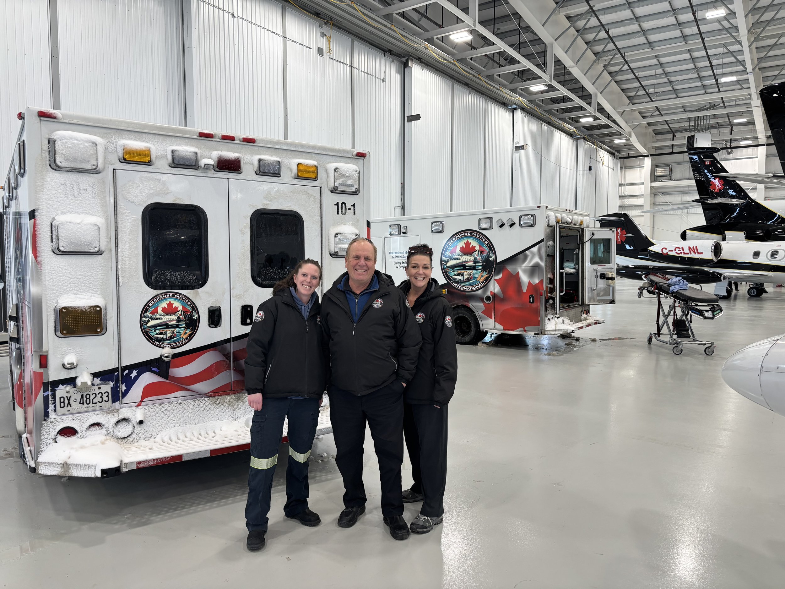 Three emergency responders standing inside an aircraft hangar with emergency response vehicles and private jets in the background. The vehicles are marked with Canadian and American symbols, indicating an international emergency response team.