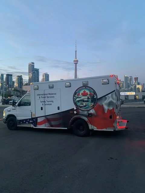 A mobile medical and travel services vehicle with Canadian and American flags painted on it, a logo, and city skyline with a tall tower in the background.