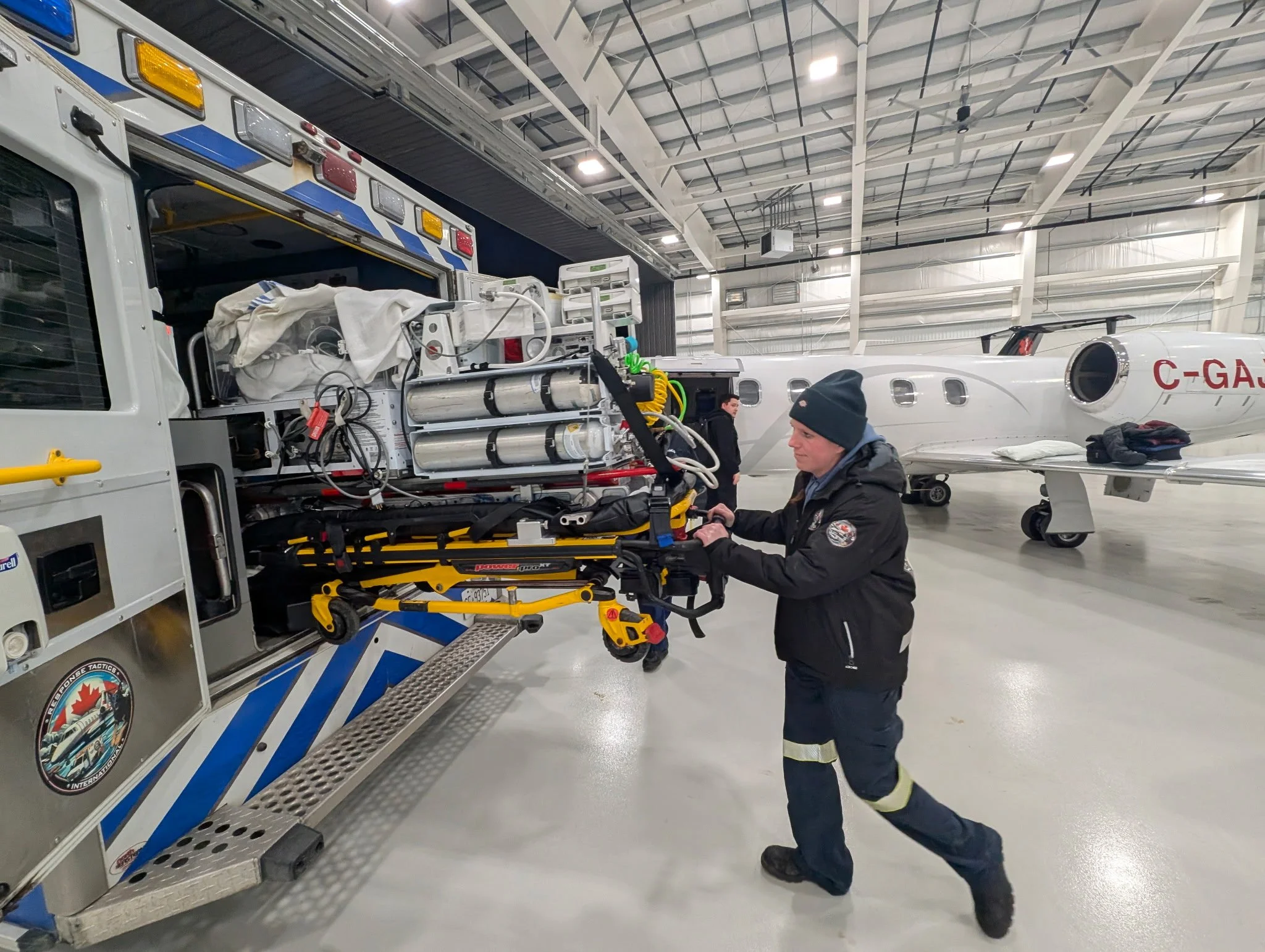 A firefighter in uniform and a beanie pushing a medical emergency stretcher with equipment into an aircraft hangar, with a private jet in the background.