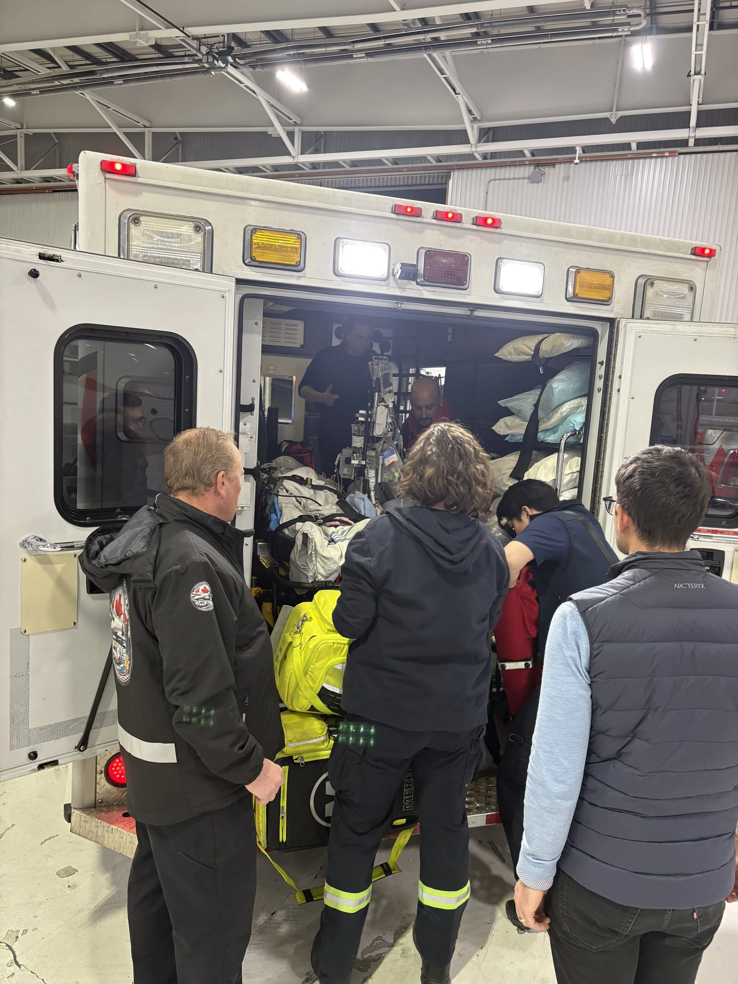 Group of people, including emergency medical personnel, gathered around the back of an ambulance inside a garage.