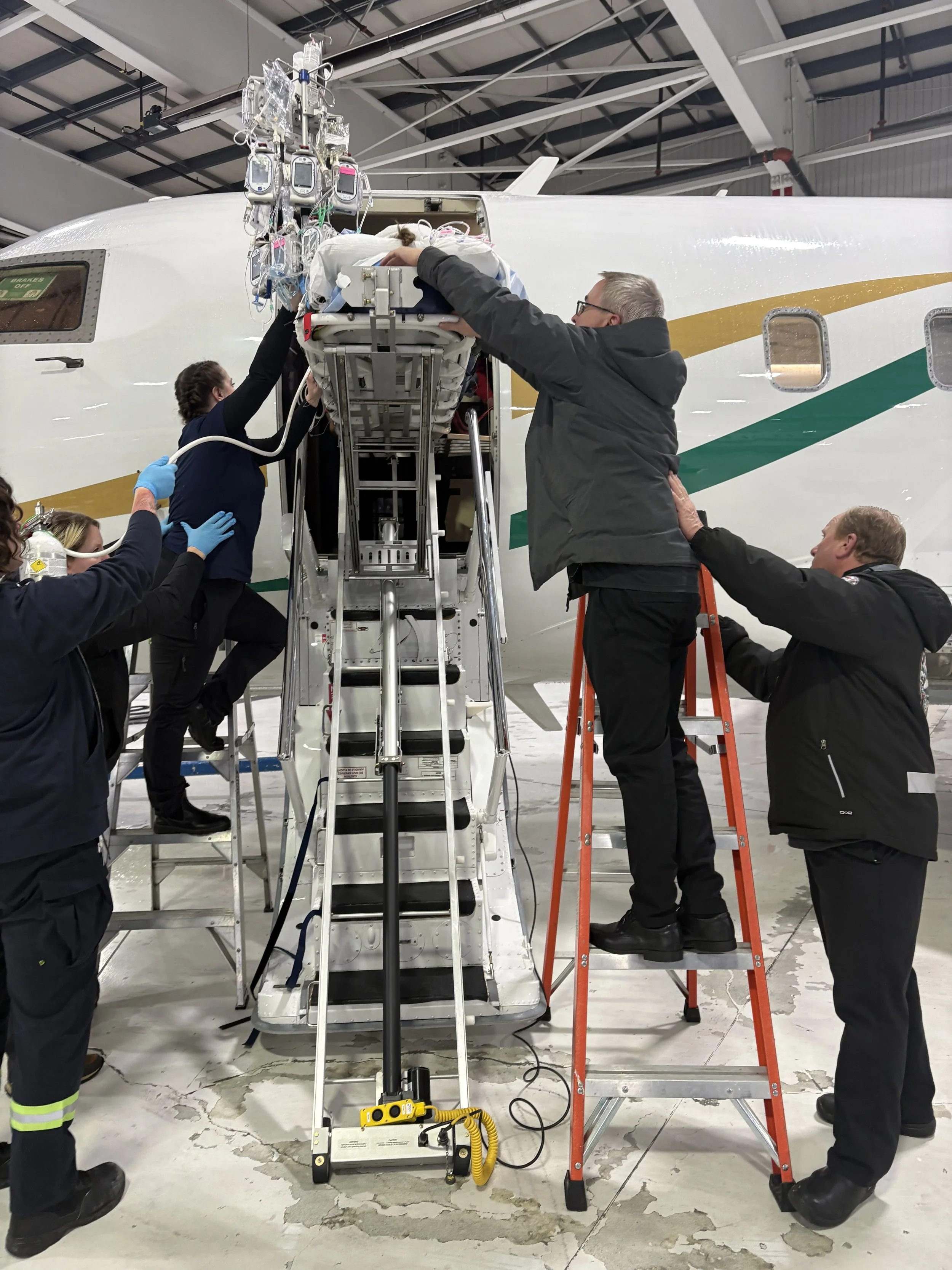 Medical personnel attending to a patient on an aircraft stairs inside a hangar, with medical equipment hanging nearby.