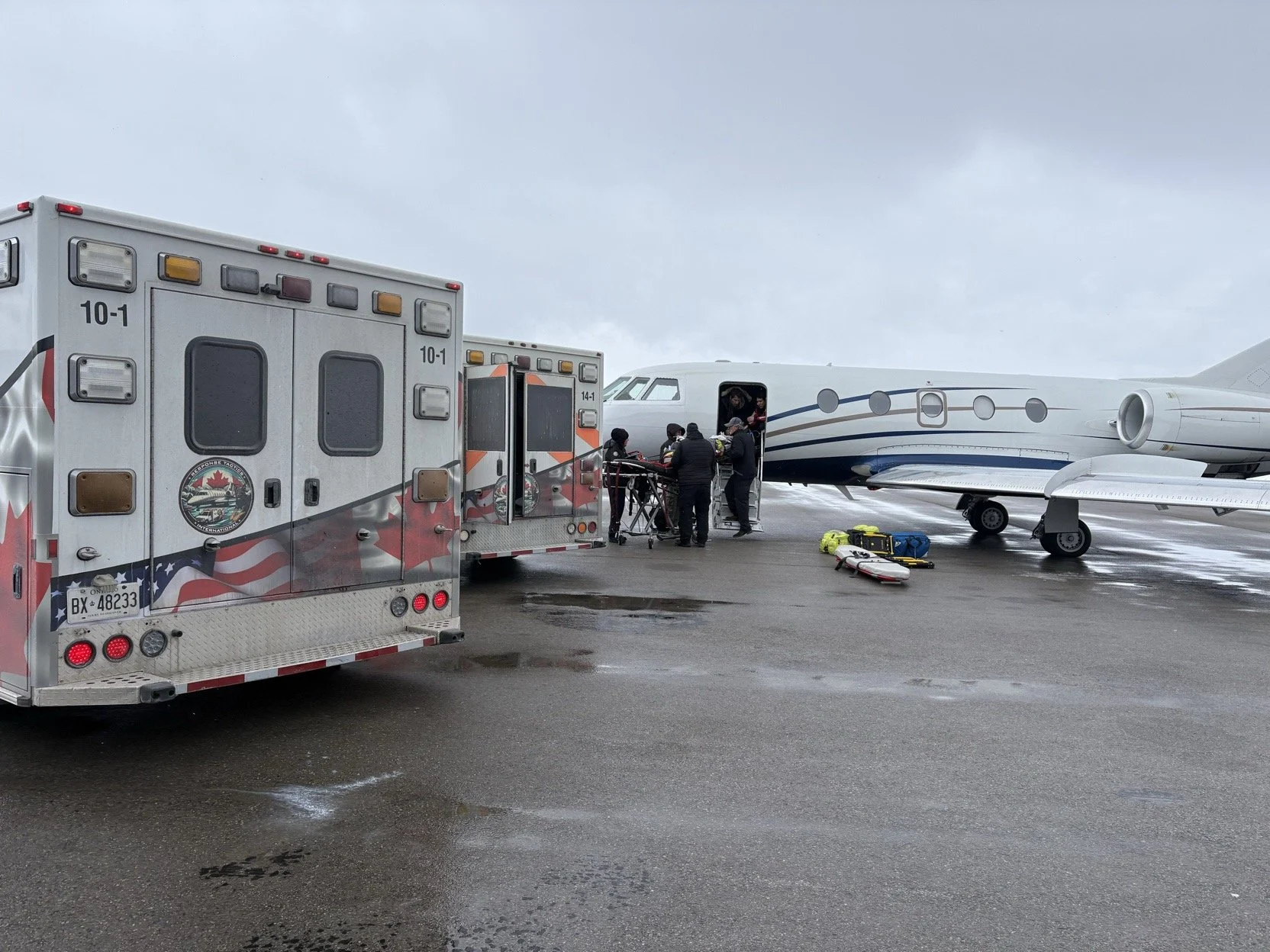 Emergency responders assist an injured person on a stretcher disembarking from a private jet on an overcast wet tarmac, with medical equipment nearby.