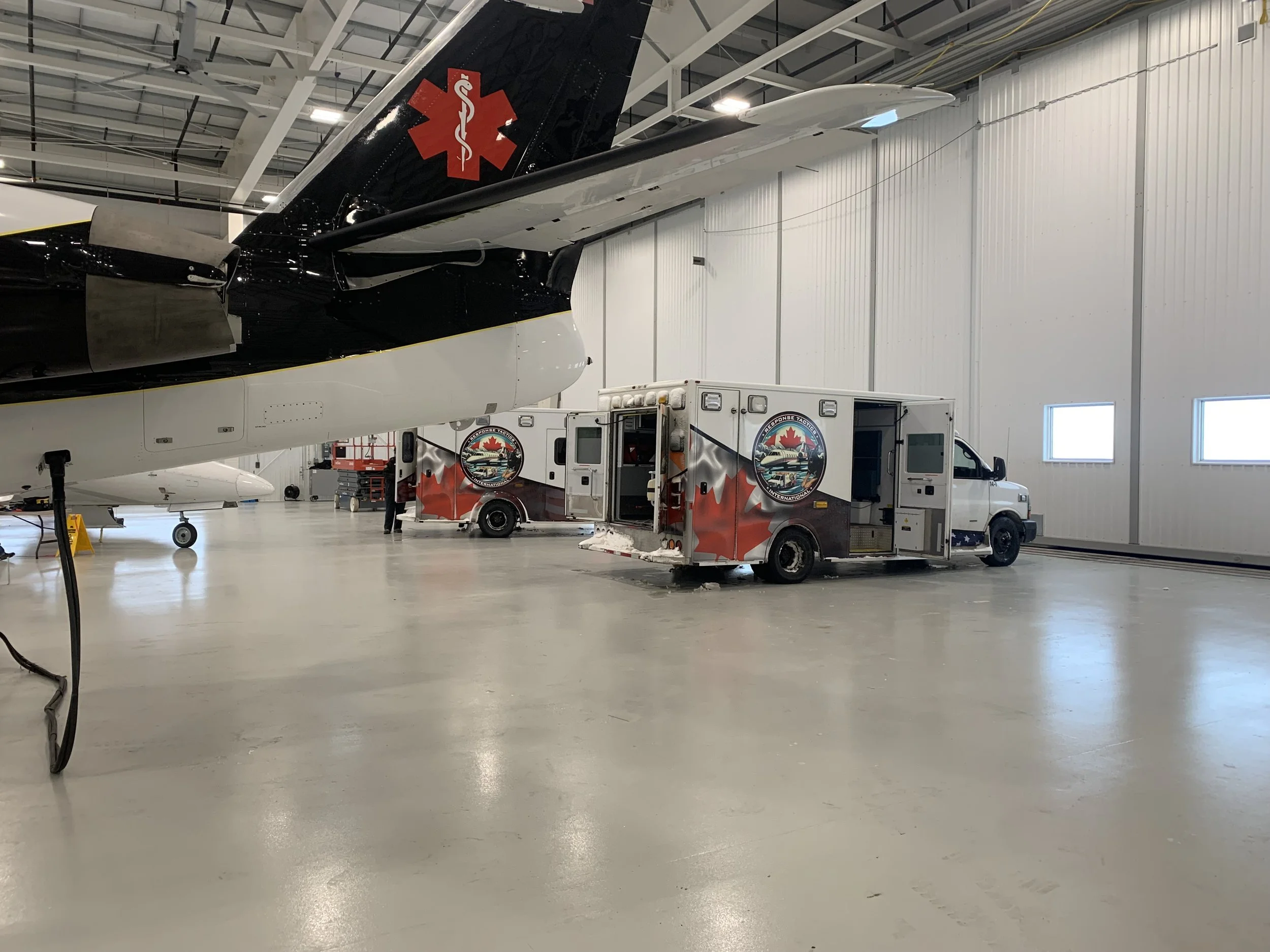 Medical emergency helicopter and ambulance inside an aircraft hangar with the Canadian flag emblem on the ambulance and an emergency symbol on the helicopter tail.