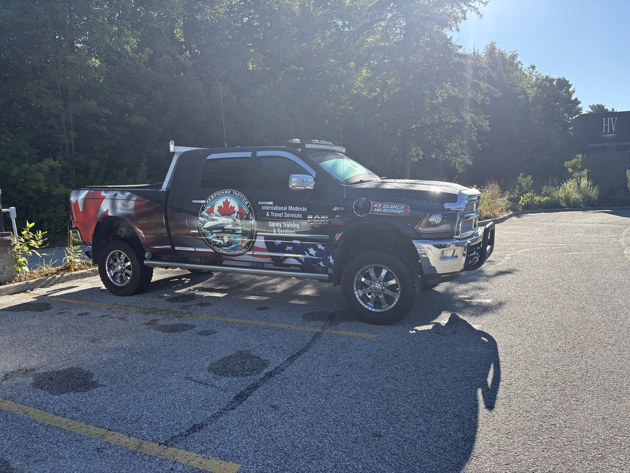 Black pickup truck with military-themed decals and logos, parked in a parking lot with shadows from nearby trees, and a sign reading 'HV' in the background.
