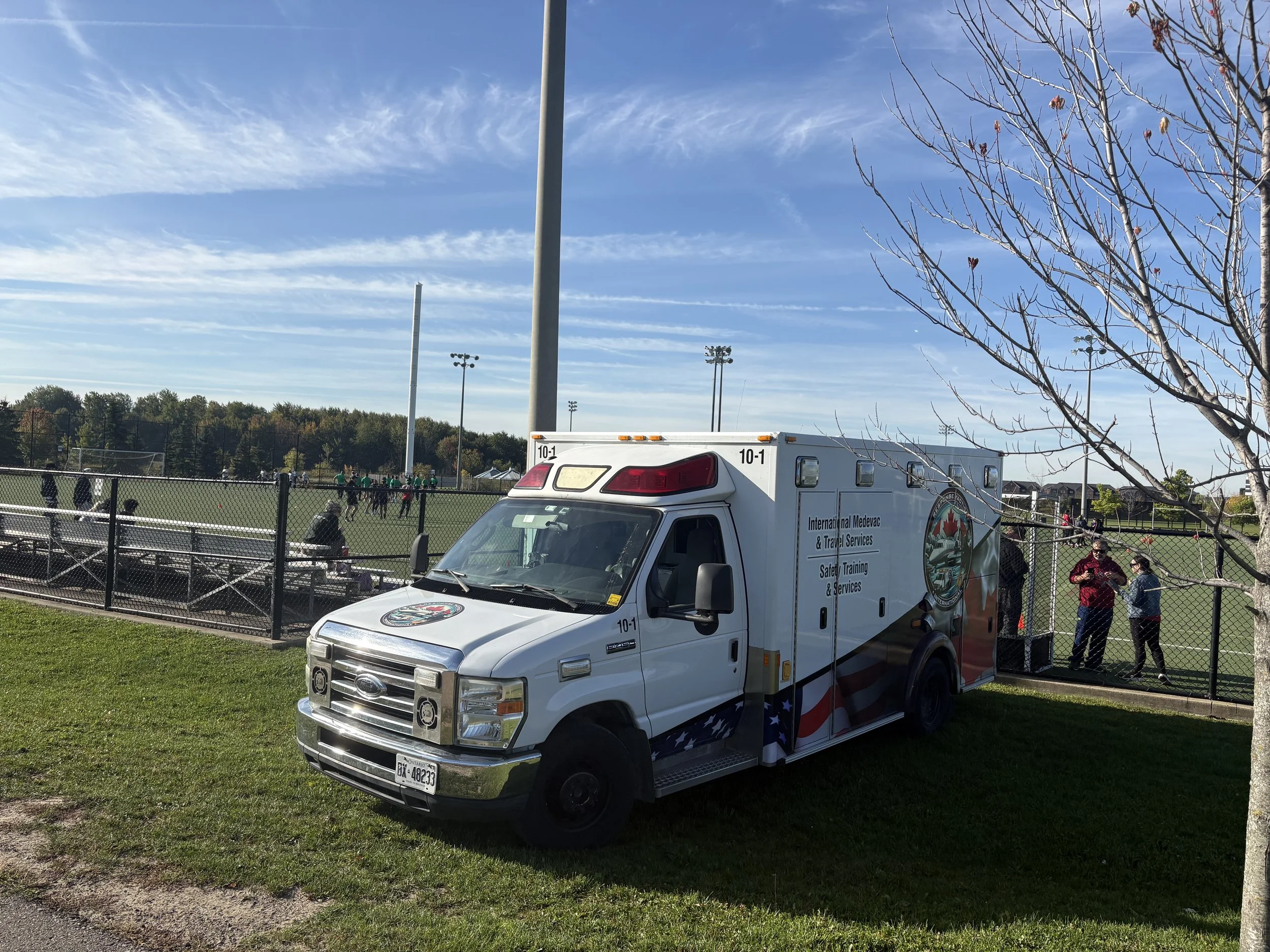 An emergency ambulance parked on grass near a football field with people playing on the field and a few people standing and talking near the fence. The sky is clear with some clouds.