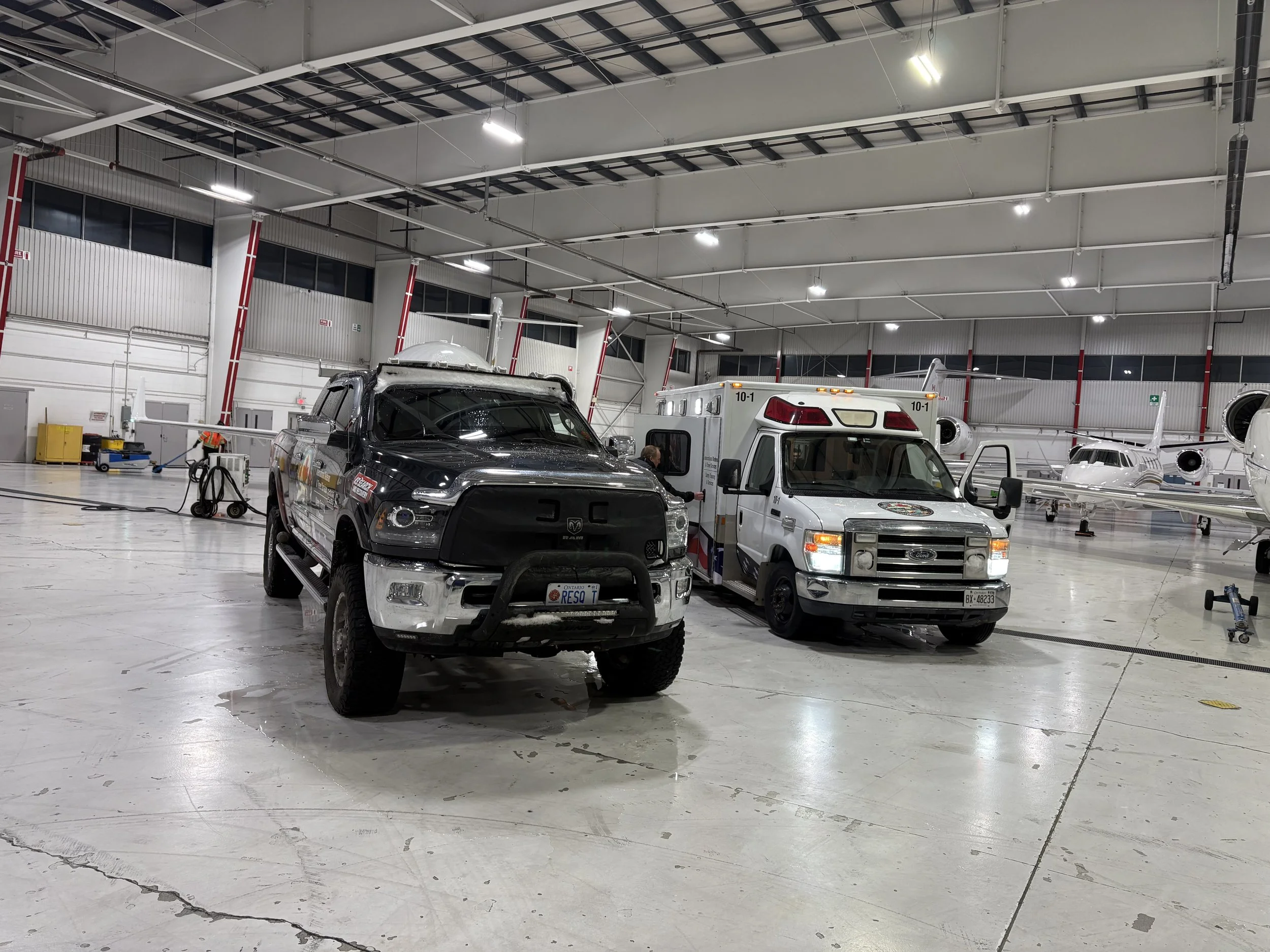 Inside an aircraft hangar with a police pickup truck and an ambulance parked, with private jets in the background.