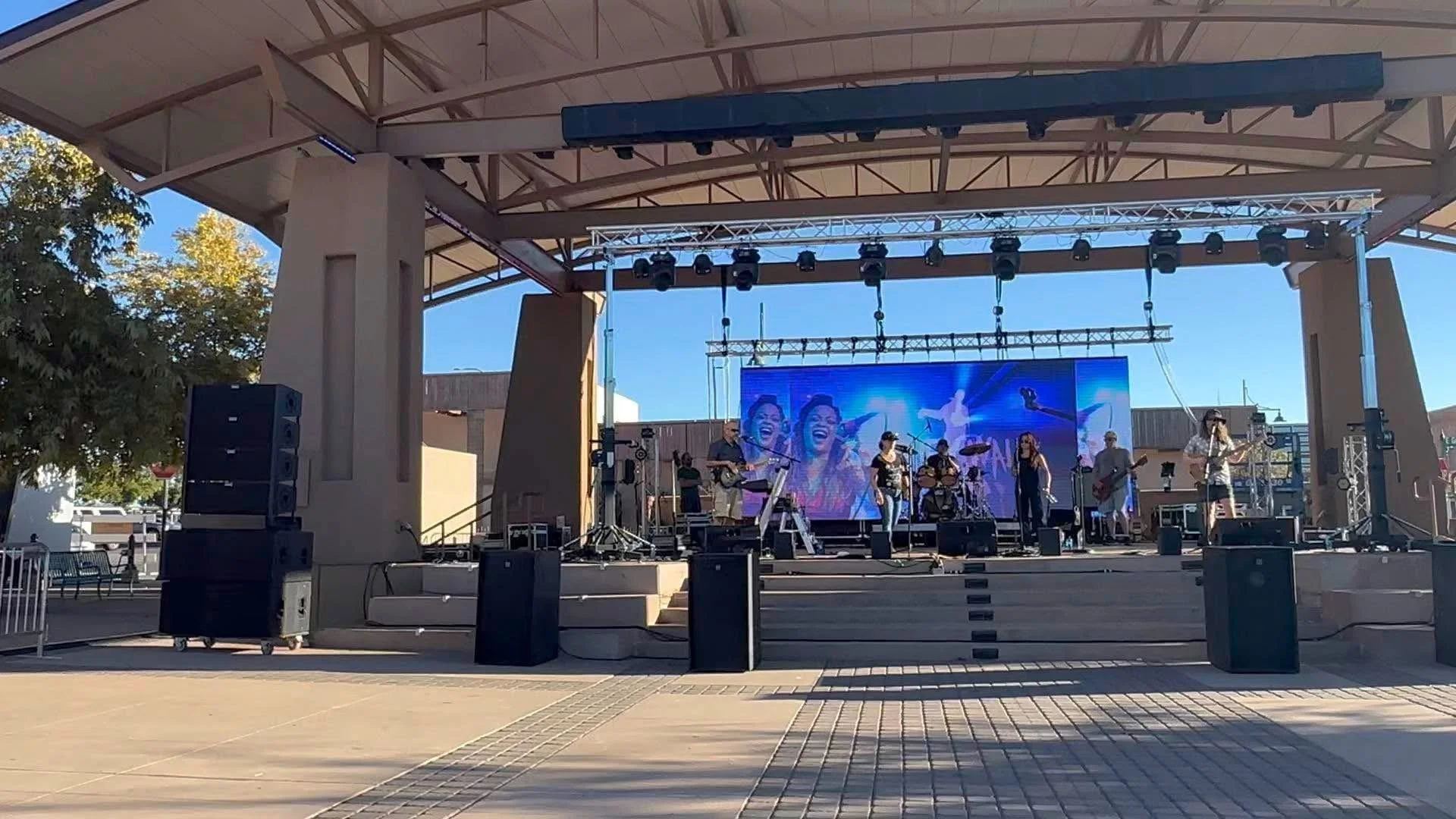 An outdoor stage with a band performing, large LED screen displaying a colorful image of smiling faces, and various musical equipment and speakers.
