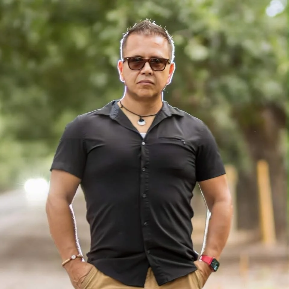 A man wearing sunglasses, a black button-up shirt, and beige pants standing outdoors on a tree-lined street.