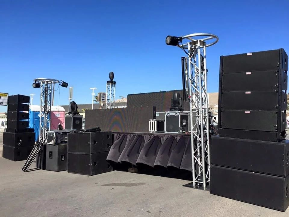 Setup of audio and lighting equipment on a stage outdoors, including speaker stacks, trussing, and a large LED screen.