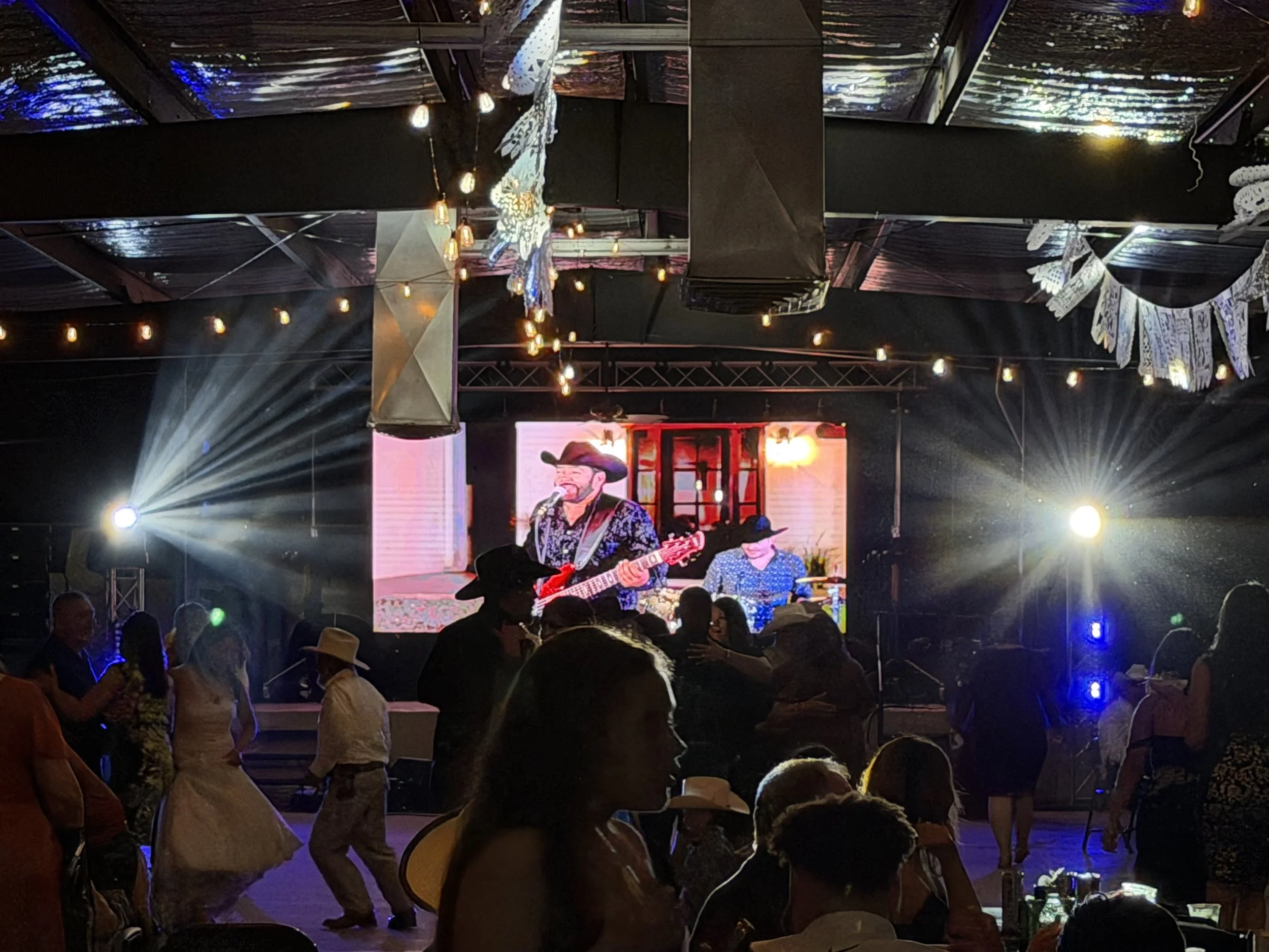 People dancing and socializing at a lively indoor event with a stage, a large screen displaying musicians, and decorative lights hanging from the ceiling.