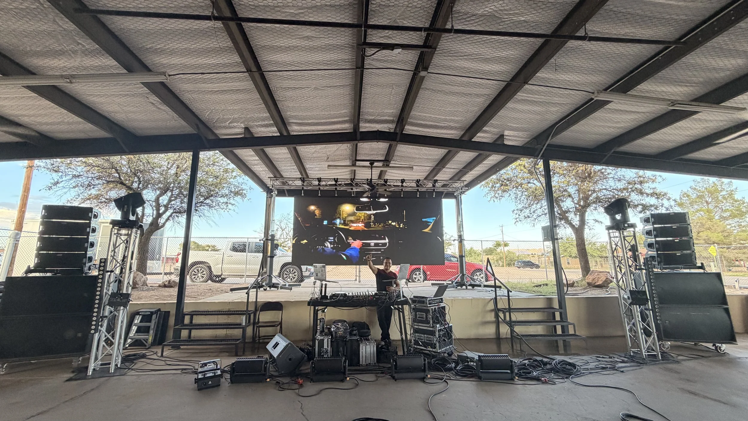 An outdoor stage setup with sound equipment, a DJ, and a large LED screen displaying a scene from inside a vehicle at night, with parked cars and trees in the background.
