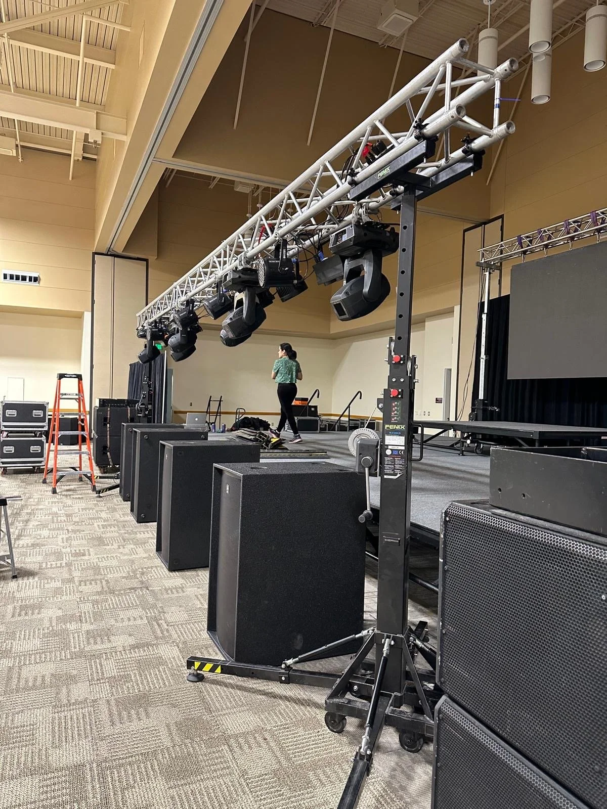 Stage setup with lighting equipment, speakers, and a person walking on stage, in a large indoor venue.