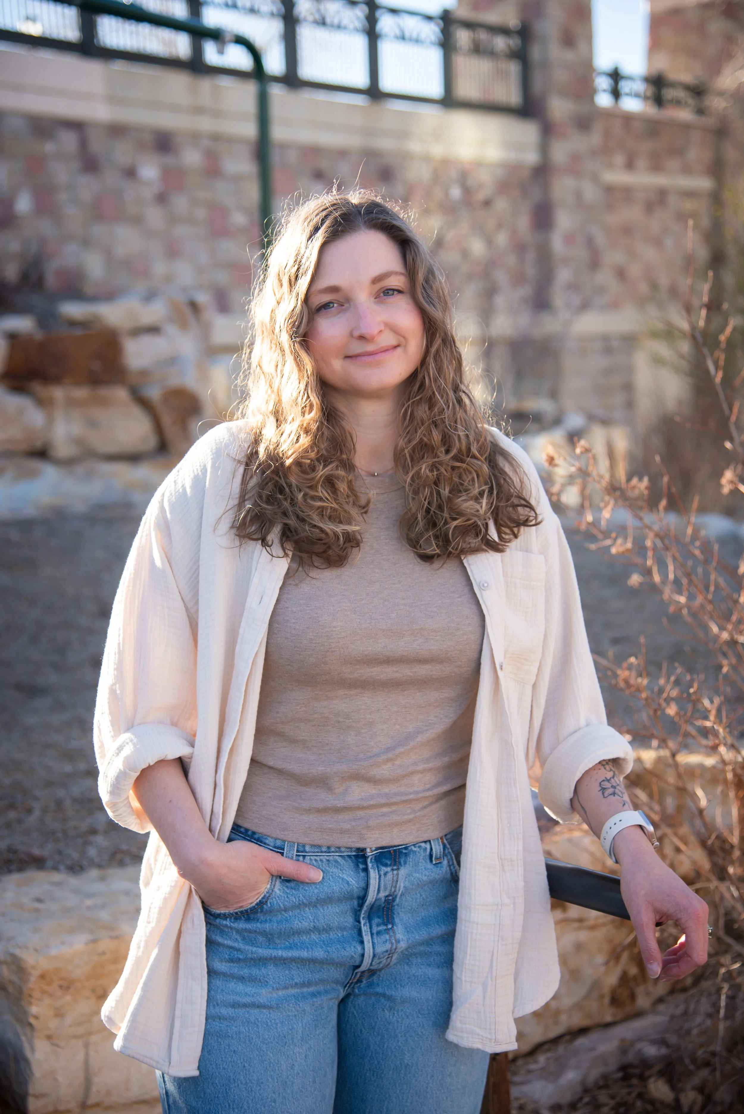 A woman with wavy hair standing outdoors, wearing a beige t-shirt, light-colored button-up shirt, and blue jeans, with her left hand resting on a railing and her right hand in her pocket.