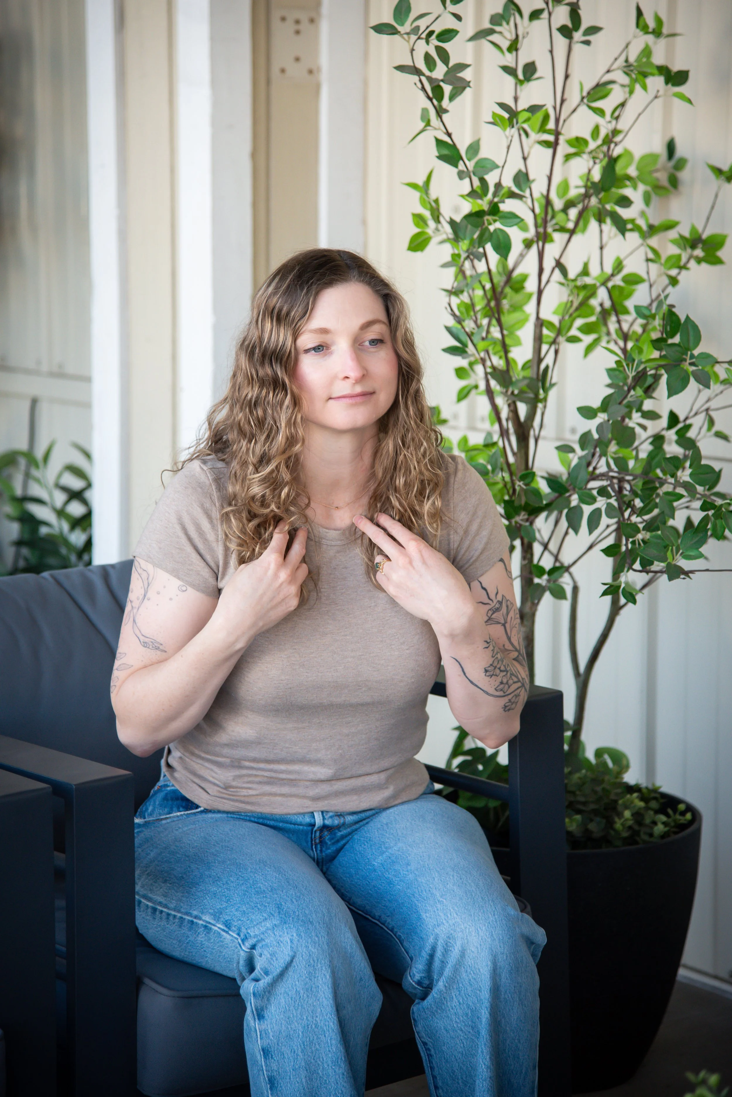 Laura Vogel with wavy brown hair and tattoos on her arms sitting on a black chair, demonstrating tapping, in front of a large leafy plant and a light-colored wall.