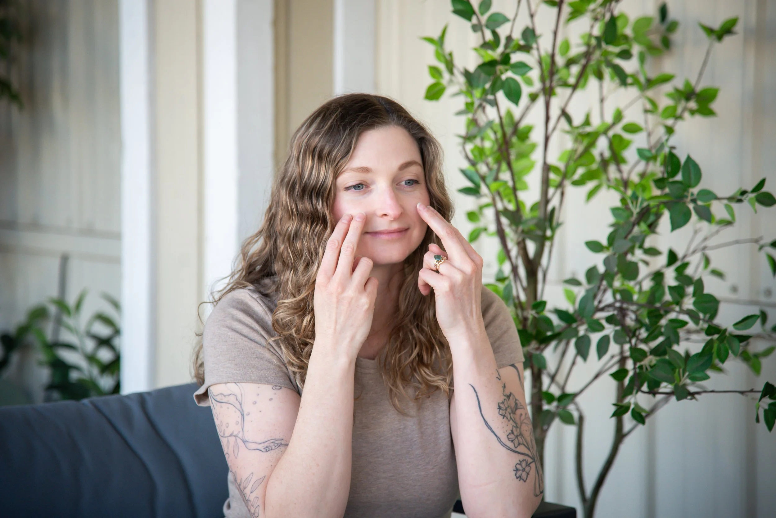 A woman with wavy brown hair and tattoos on her arms is sitting indoors, touching her face near her cheeks, with a slight smile, and a large plant is behind her.