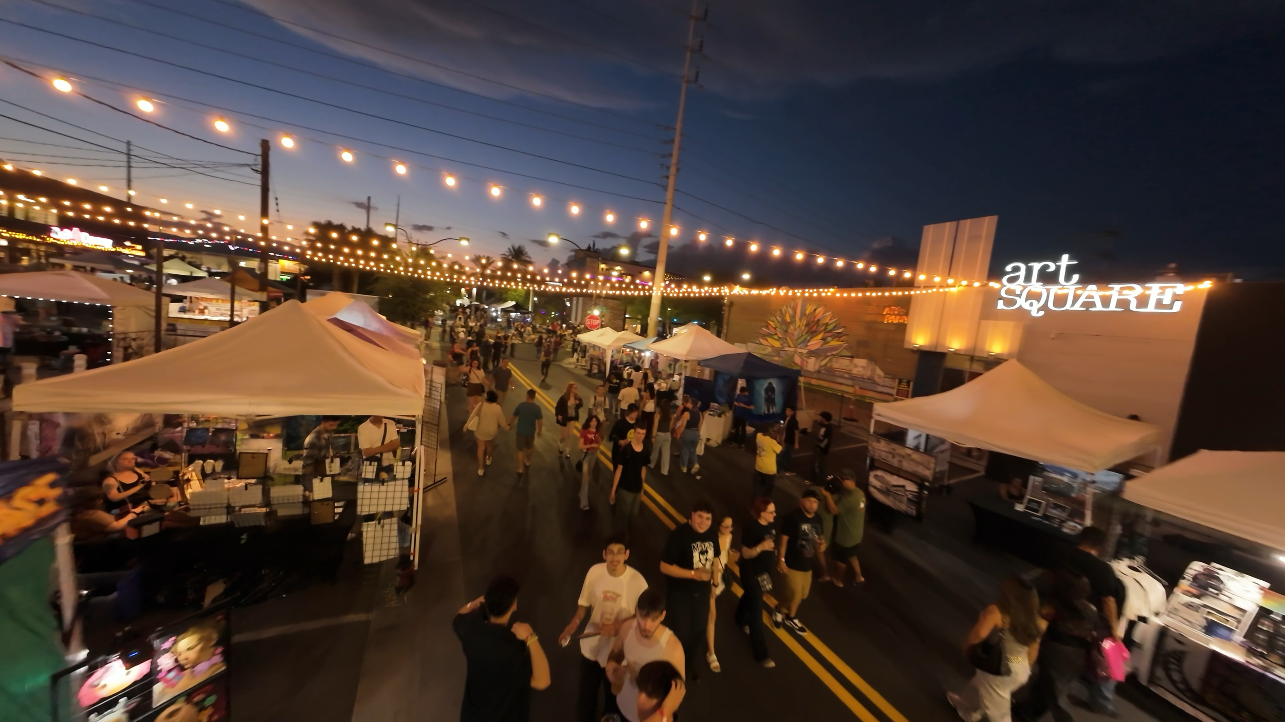 Nighttime street fair at Art Square with string lights, tents, and people walking and shopping during First Friday.