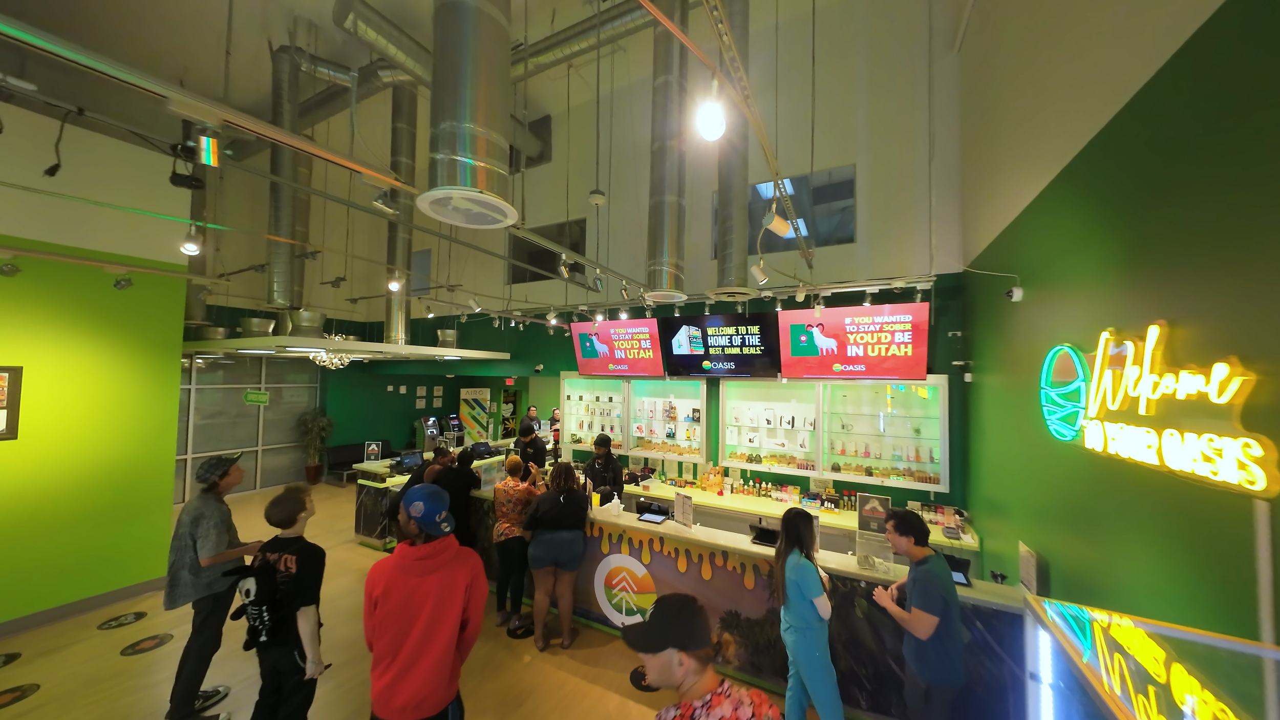 Inside a busy dispensary store with people lining up at the counter, colorful neon signs, digital screens displaying advertisements, and a vibrant green wall with a neon logo.
