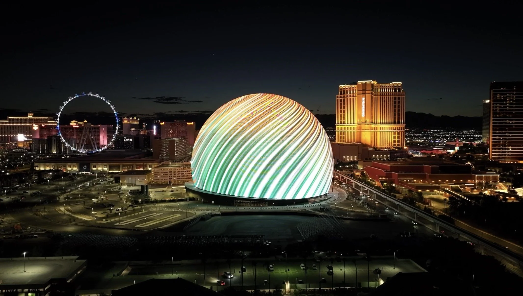 Nighttime cityscape with illuminated buildings, a large domed structure with rainbow-colored lights, and a Ferris wheel in the background.