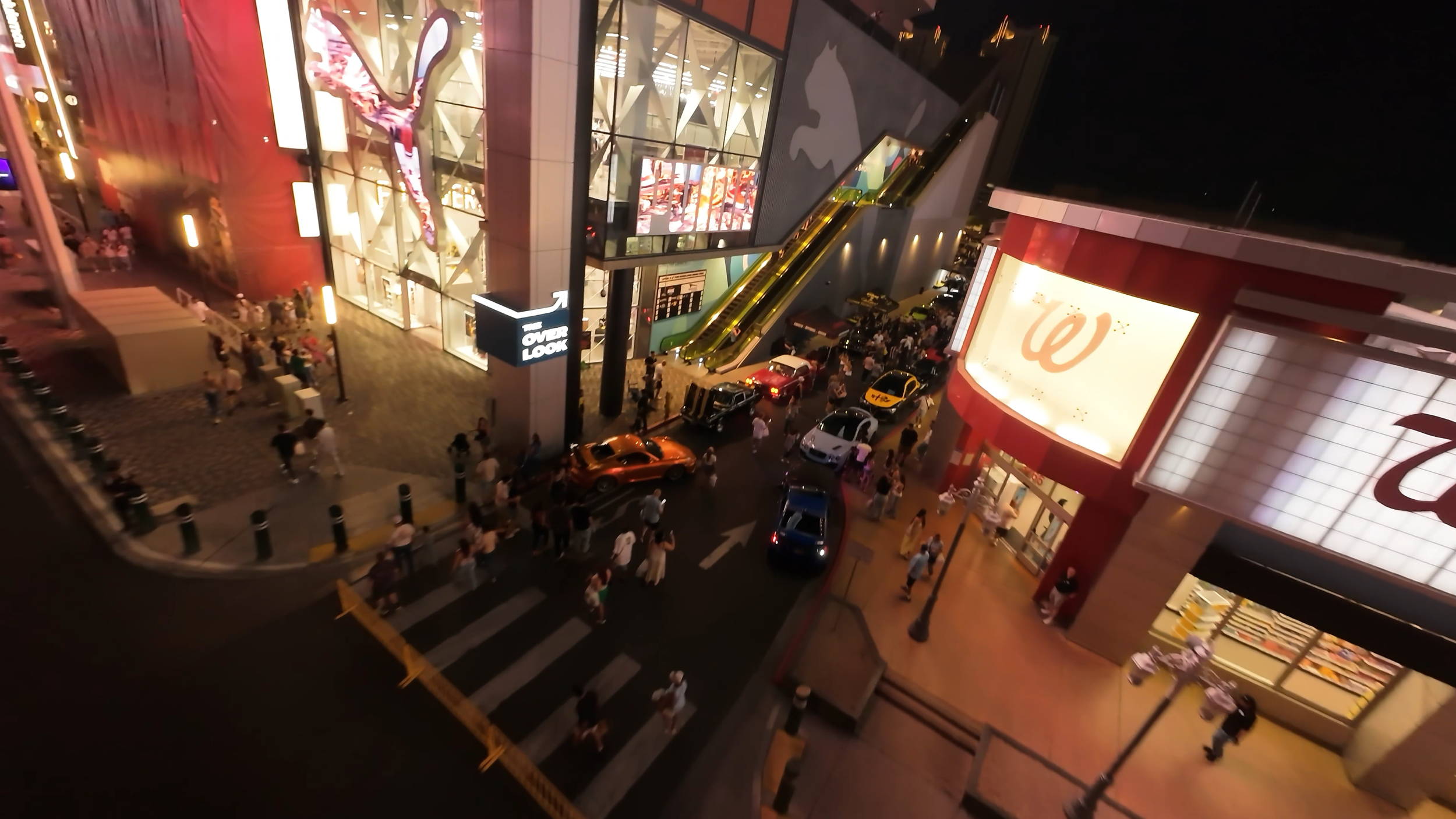 Nighttime view of a busy shopping mall entrance with people walking, cars parked, bright signage, and escalators visible.