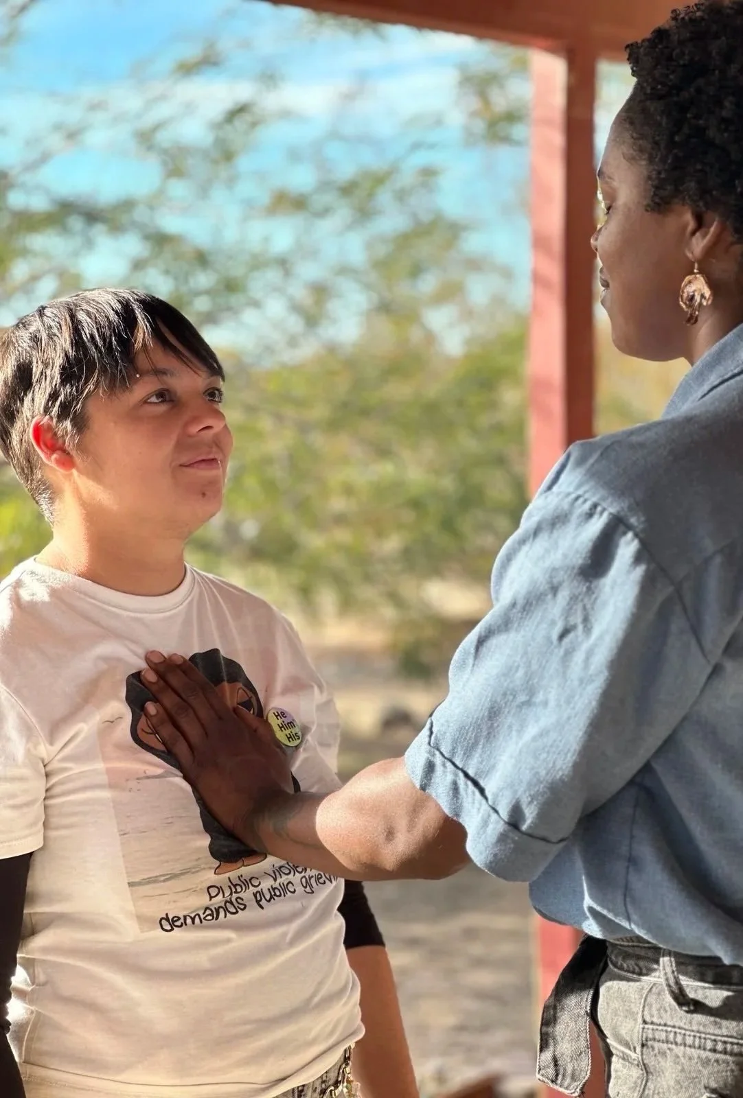 A Black person with short curly hair and earrings is placing their hand on a shorter Latinx person's chest. The shorter person is looking up with a focused expression. They are outside with trees and a blue sky in the background.