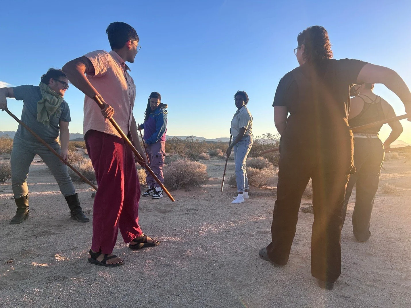 A multiracial group of people standing in a desert landscape during sunset, holding jos, practicing with each other.