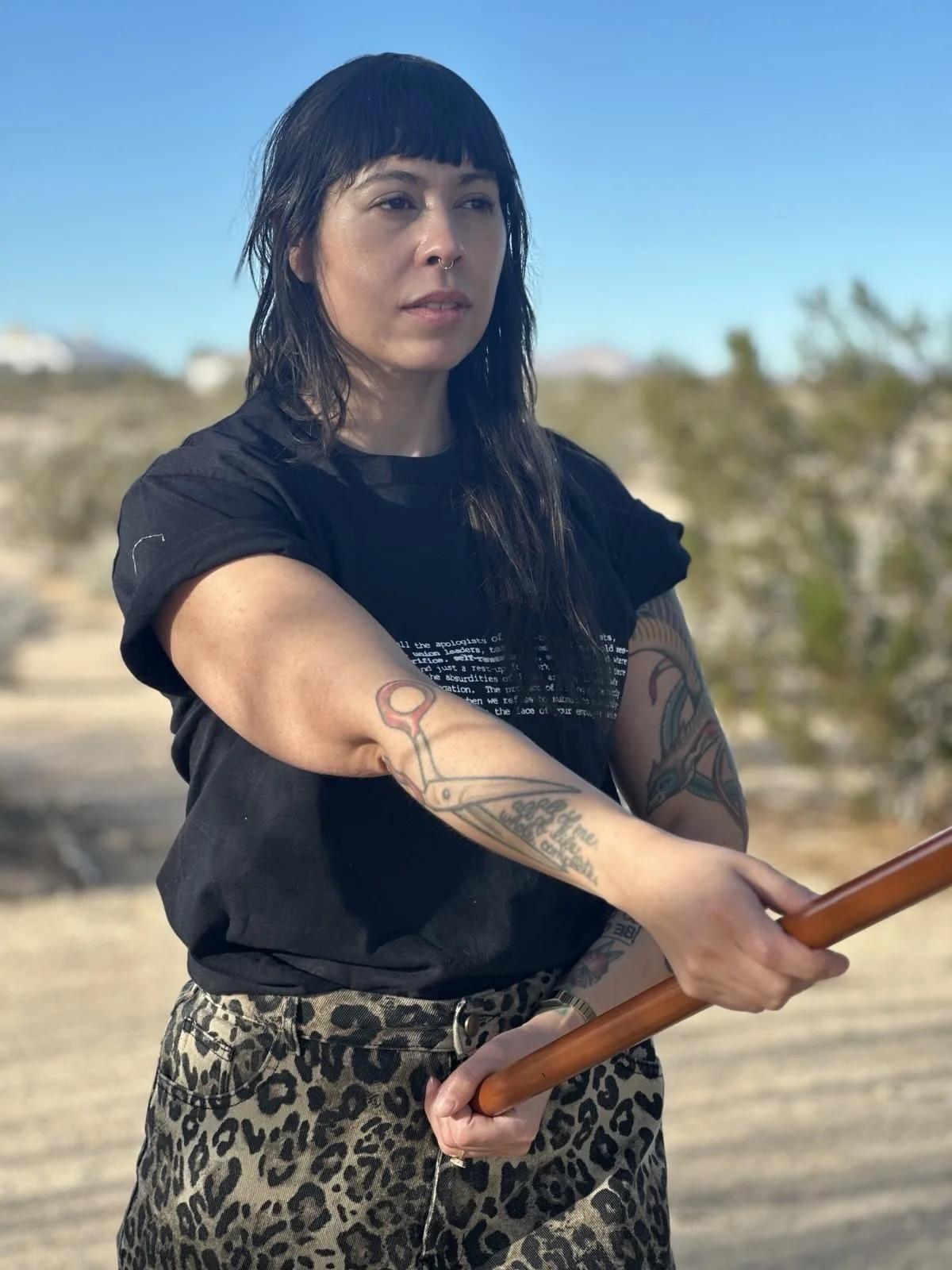 B Stepp, a Black person with black hair, tattoos, and a nose piercing holding a jo outdoors, with desert landscape and clear blue sky in the background.