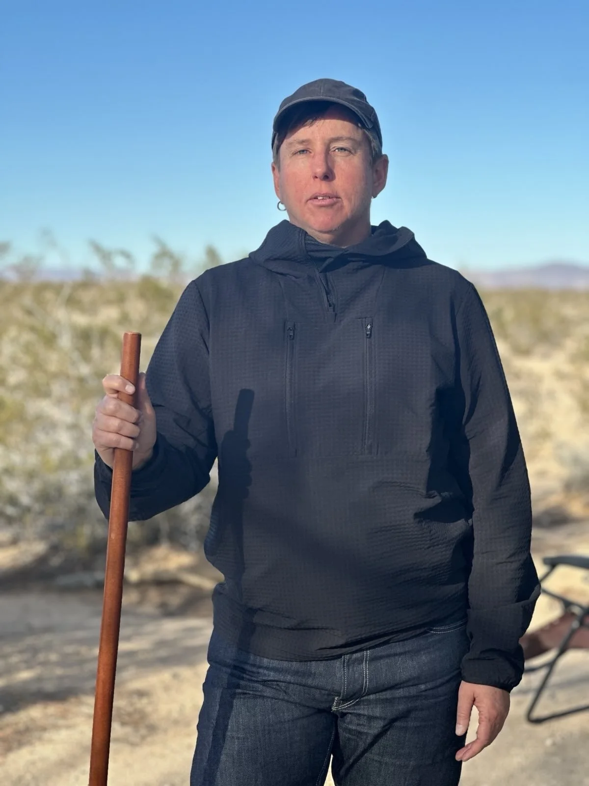 Ream, a white person standing outdoors in a desert landscape, wearing a hoodie and baseball cap, holding a jo, with a clear blue sky and sparse desert vegetation in the background.